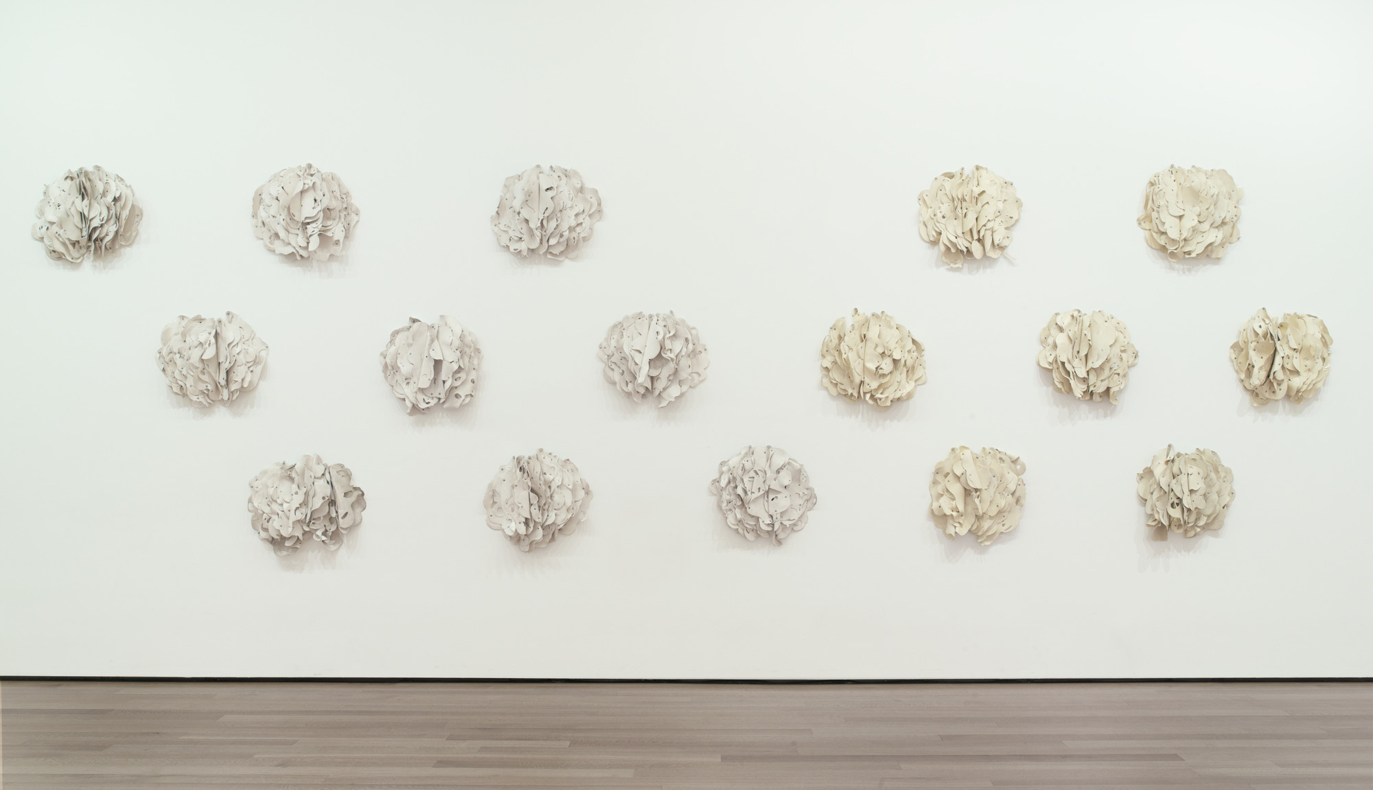 The image shows a white wall with fifteen round, crumpled paper-like structures in various shades of white and beige, arranged in a grid pattern. The floor is light brown wood. This is an AI generated description. To give feedback, email ai@moma.org.