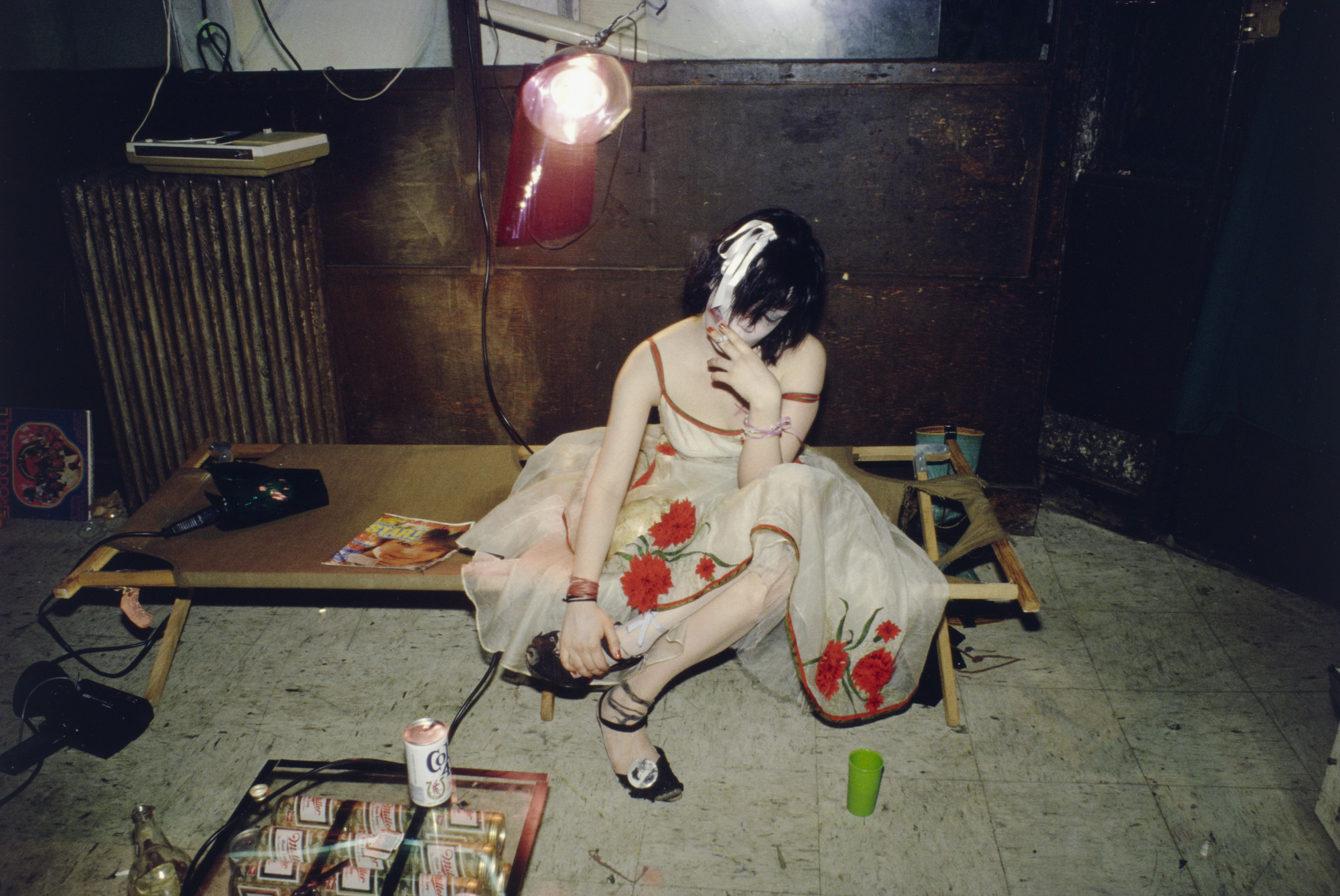 A person in a white dress with red flowers sits on a broken cot in a dimly lit room, surrounded by scattered items including a green cup and beer cans. A red light bulb hangs above, and a cast iron radiator is in the background. This is an AI generated description. To give feedback, email ai@moma.org.