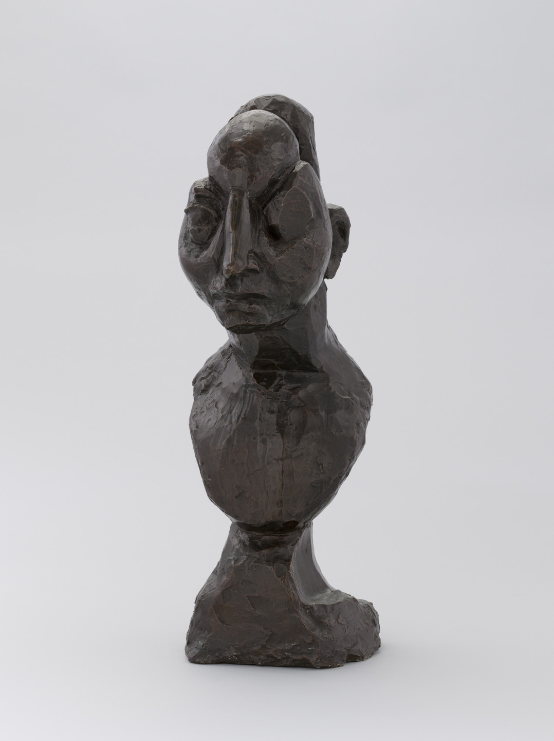 A dark, roughly textured sculpture depicts a humanoid figure with exaggerated facial features and a prominent nose. The sculpture is set against a plain light gray background. This is an AI generated description. To give feedback, email ai@moma.org.