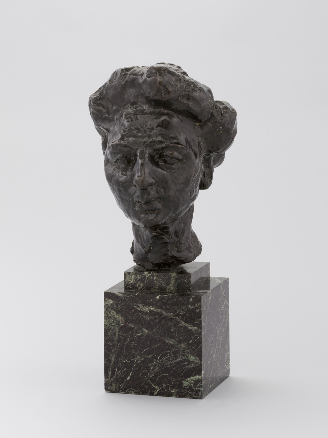 The image shows a dark brown, textured sculpture of a human head mounted on a greenish-black marble base. The background is plain and light gray. This is an AI generated description. To give feedback, email ai@moma.org.