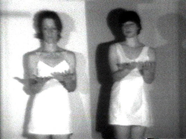Two figures in sleeveless white outfits stand side by side against a background with both light and dark areas. Each figure has their arms extended with palms up, and their blurred shadows are visible behind them. This is an AI generated description. To give feedback, email ai@moma.org.