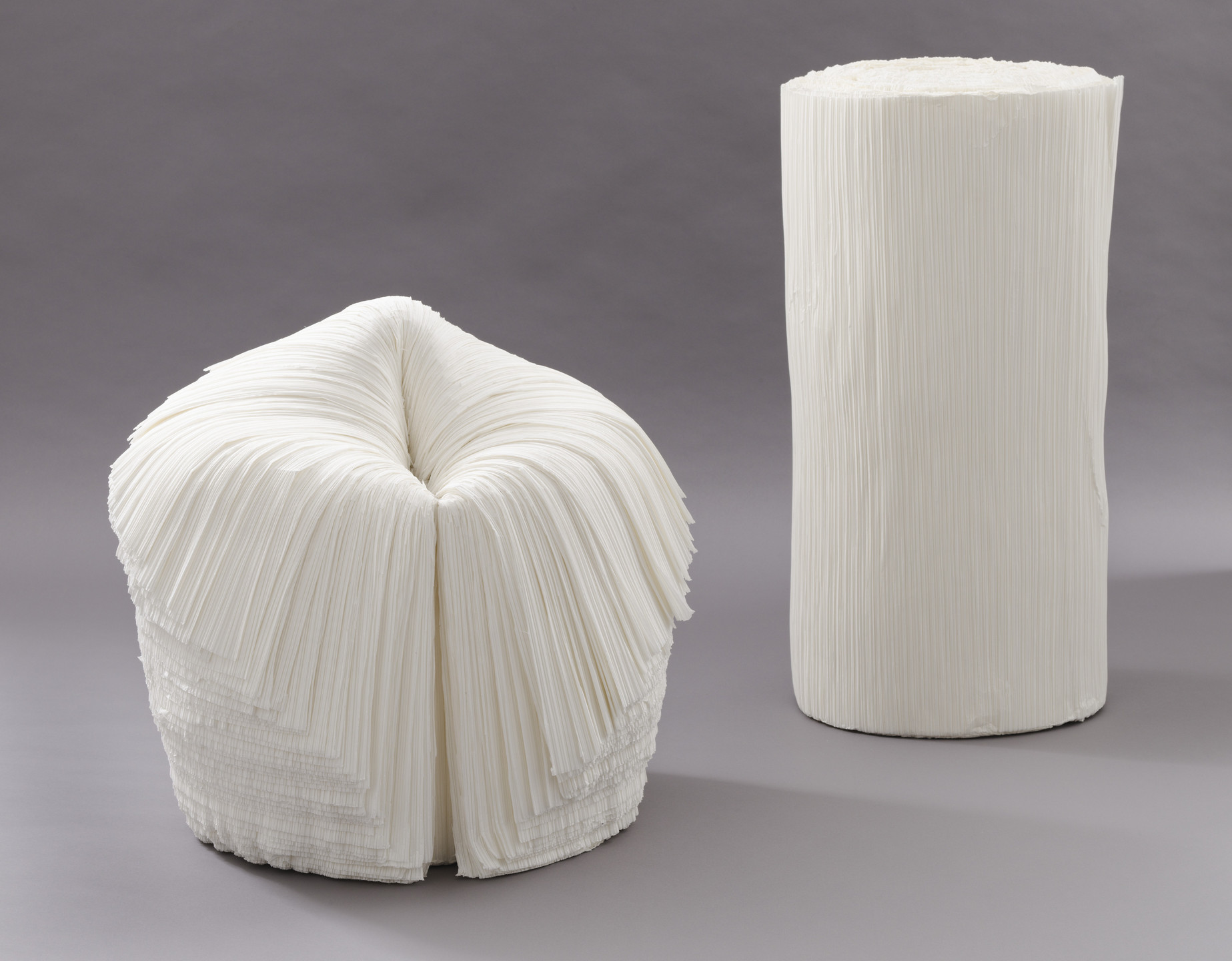 The image shows two white sculptures with a layered texture, resembling folded paper. One sculpture is squat and rounded, while the other is taller and cylindrical, both on a plain gray background. This is an AI generated description. To give feedback, email ai@moma.org.