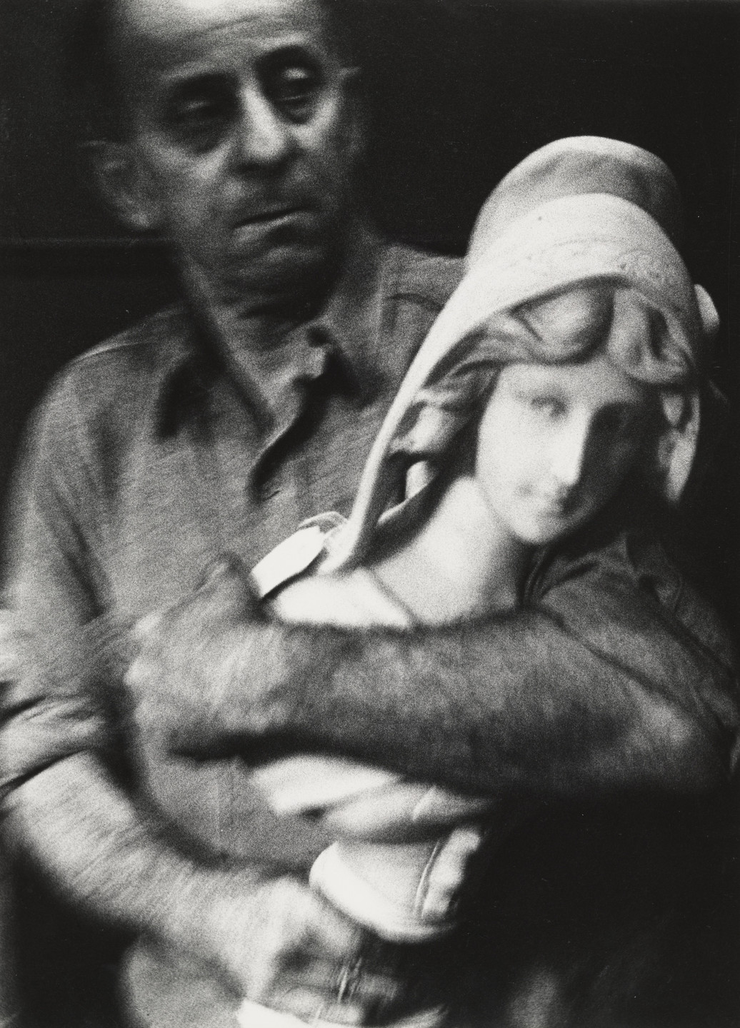A person with a serious expression holds a bust of a figure with curled hair and a head covering. The image is in black and white, with blurry details and a shadowy background. This is an AI generated description. To give feedback, email ai@moma.org.
