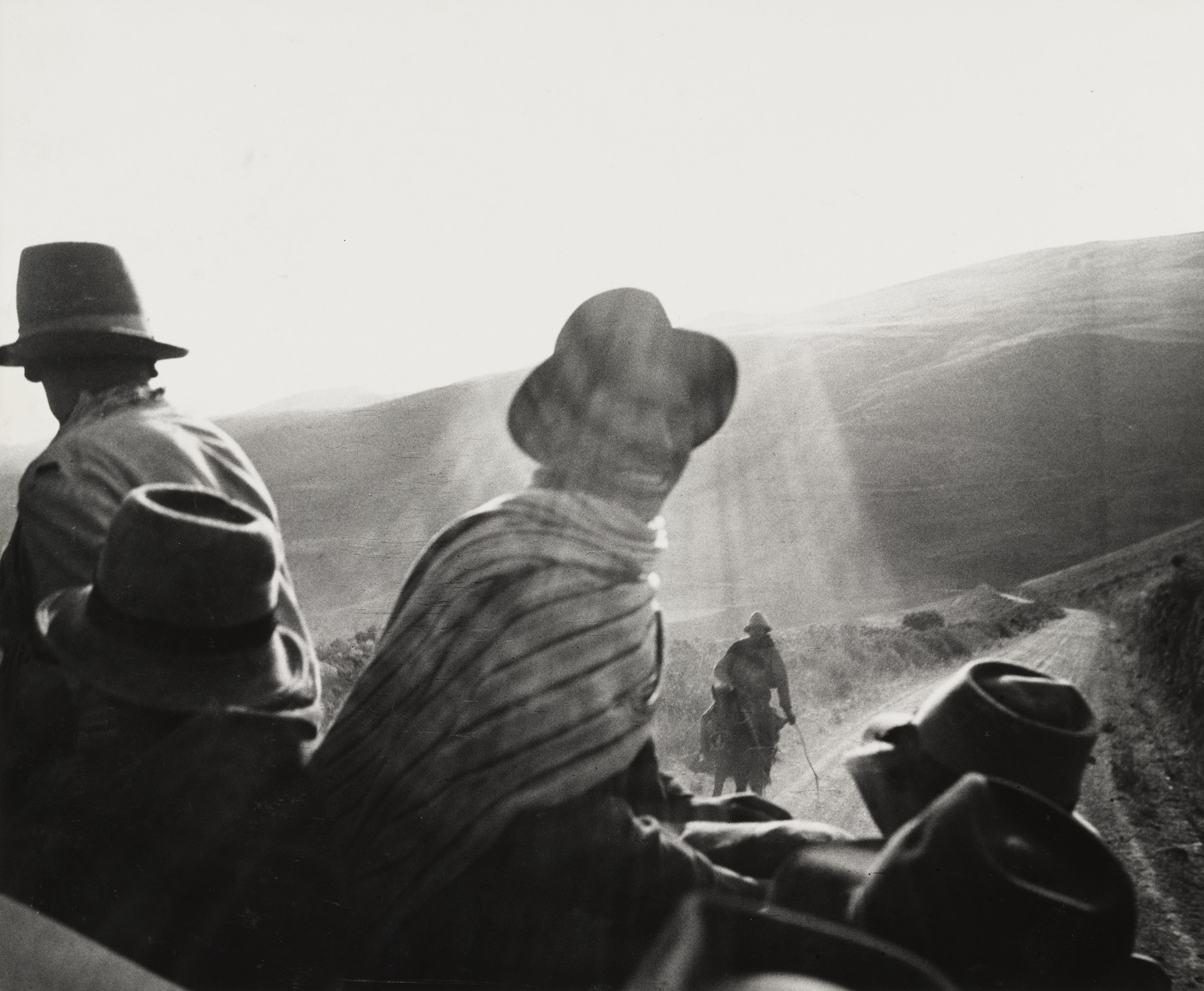 The image shows several people with hats and cloaks in the foreground, with one person looking back and smiling and rays of sunlight illuminating a rural dirt road. In the background, there is a person riding a horse amidst rolling hills. This is an AI generated description. To give feedback, email ai@moma.org.