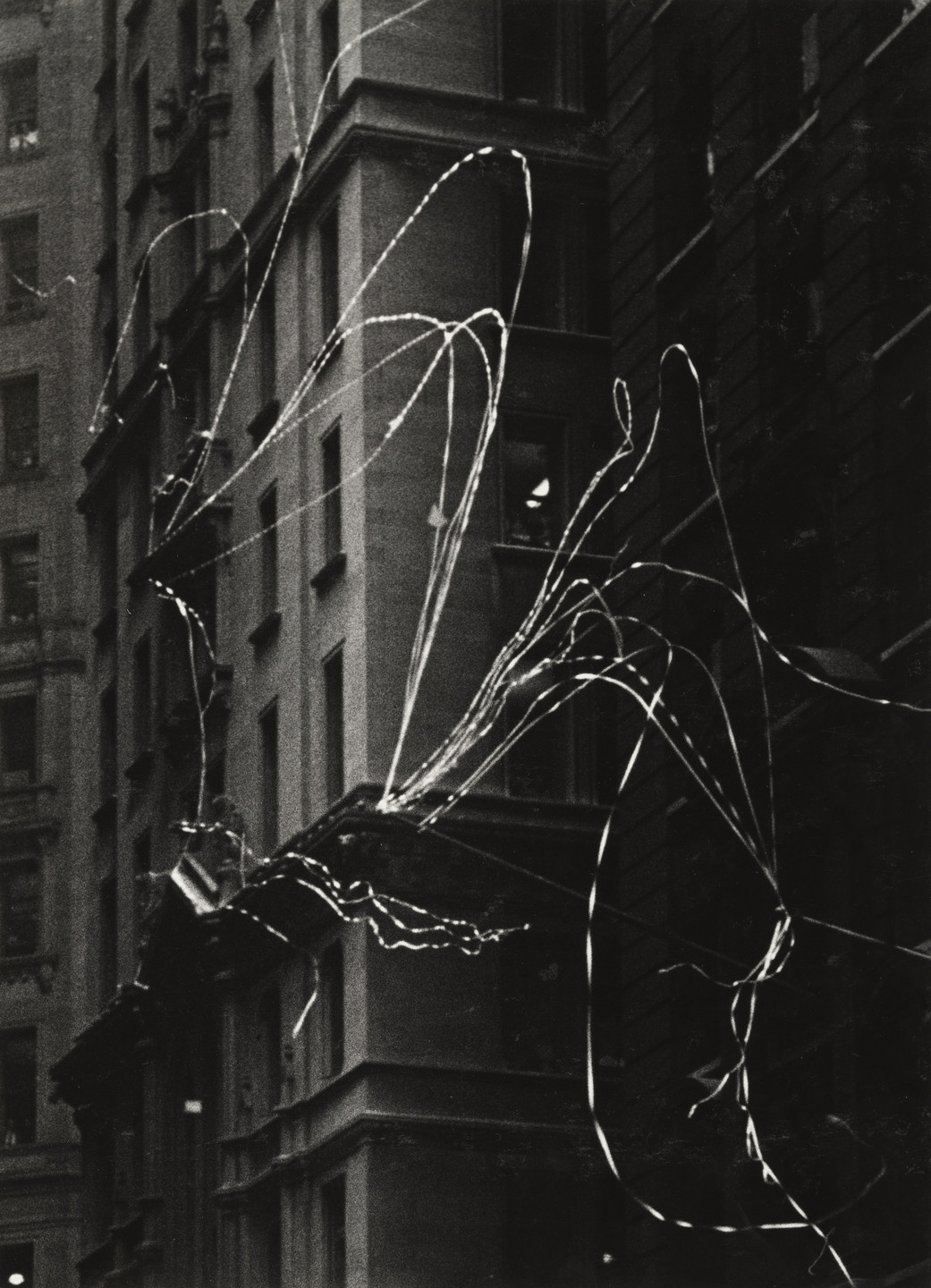 A black and white image showing tall buildings in the background with long, flowing white ribbons streaming across the scene. The buildings are stacked with windows and have intricate architectural details. This is an AI generated description. To give feedback, email ai@moma.org.