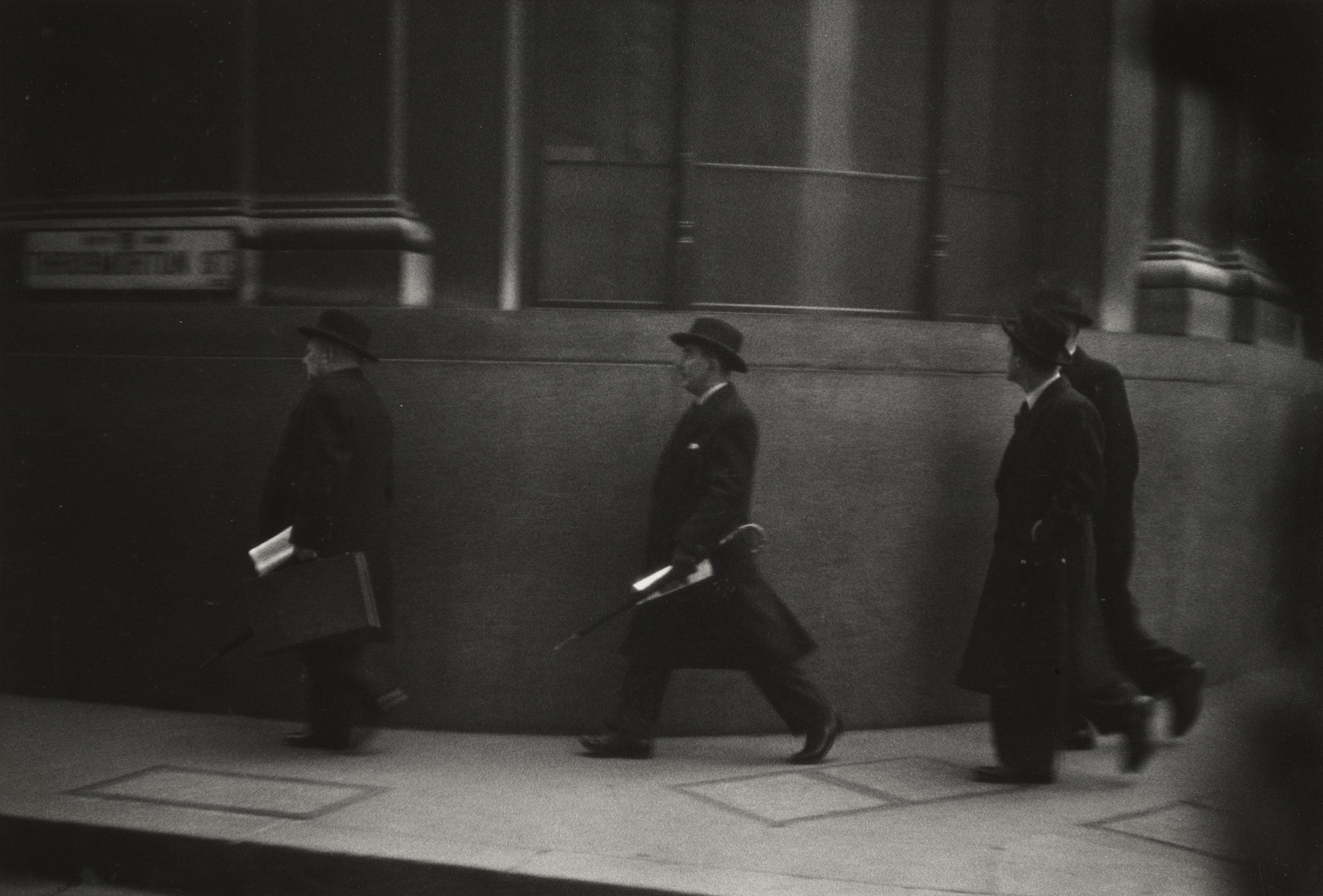 Three people wearing dark coats and hats walk in a single-file line on a sidewalk beside a dark building wall. One person carries a newspaper and briefcase, while another holds an umbrella. This is an AI generated description. To give feedback, email ai@moma.org.