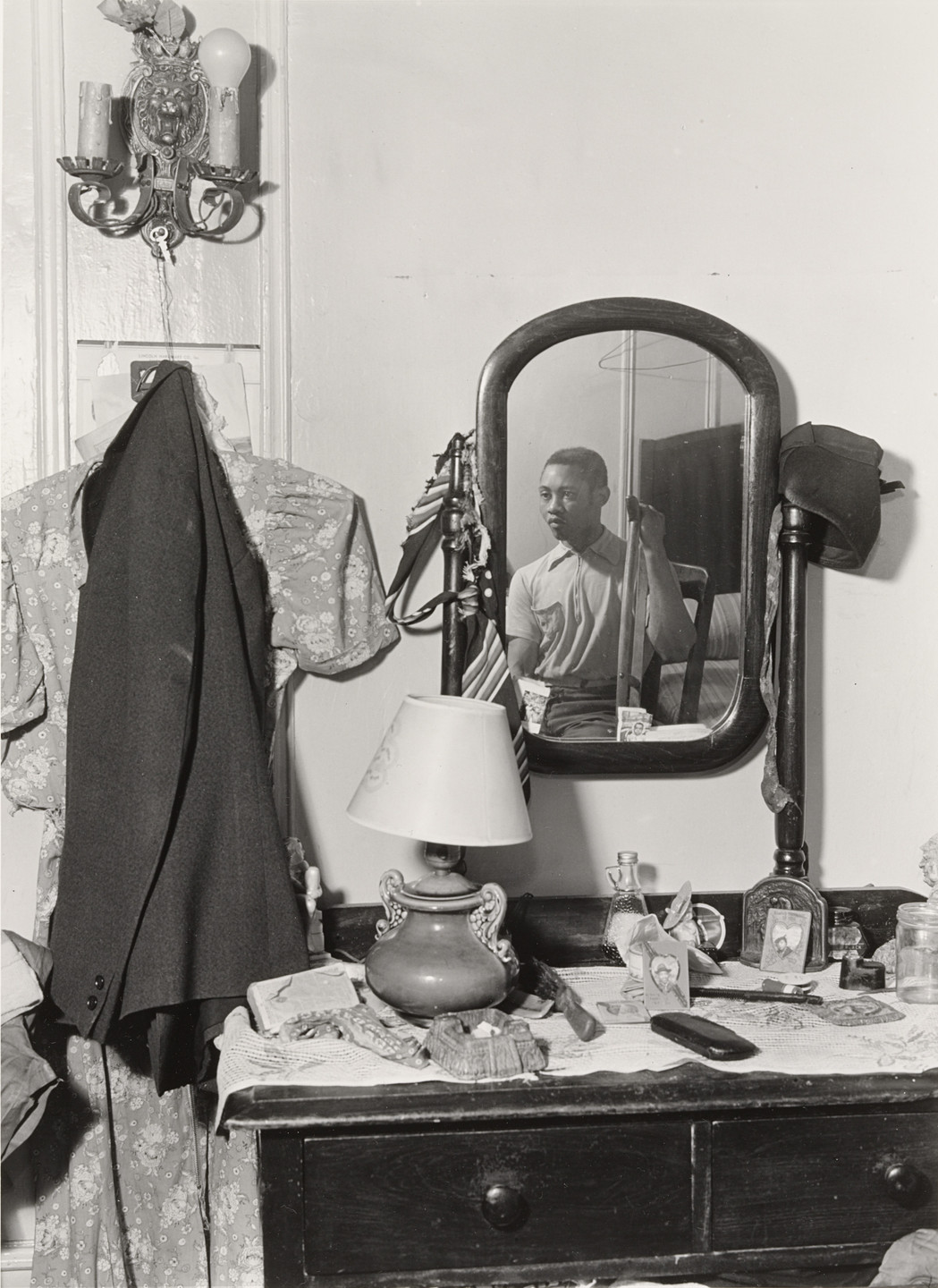 A person is reflected in an oval mirror above a dresser with various objects, including framed photos, a lamp, and a hat. A floral dress and dark jacket hang beside the mirror. This is an AI generated description. To give feedback, email ai@moma.org.