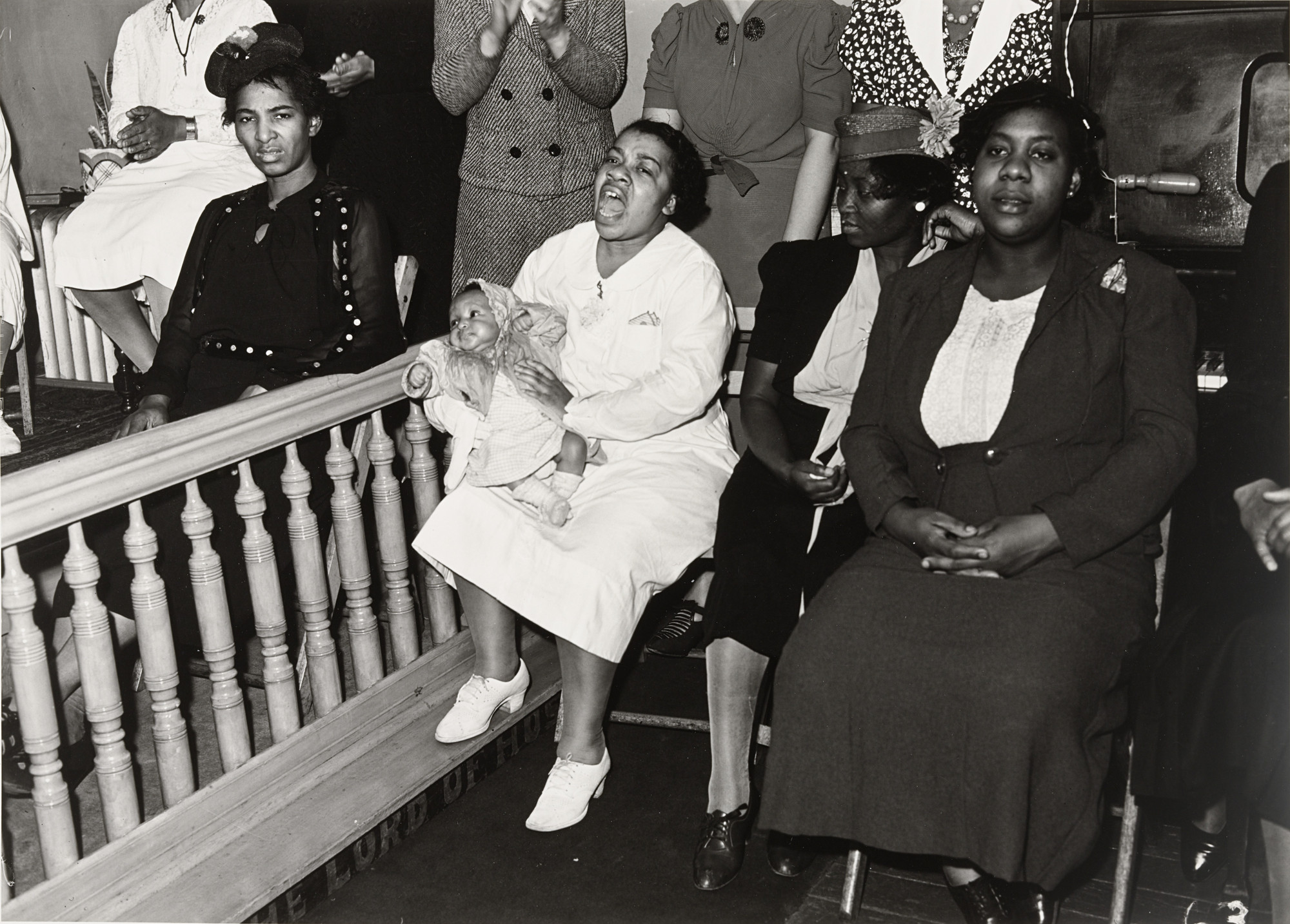 Several people are seated and standing, with some clapping. One person is holding an infant and appears to be singing or speaking passionately; the image is in black and white. This is an AI generated description. To give feedback, email ai@moma.org.