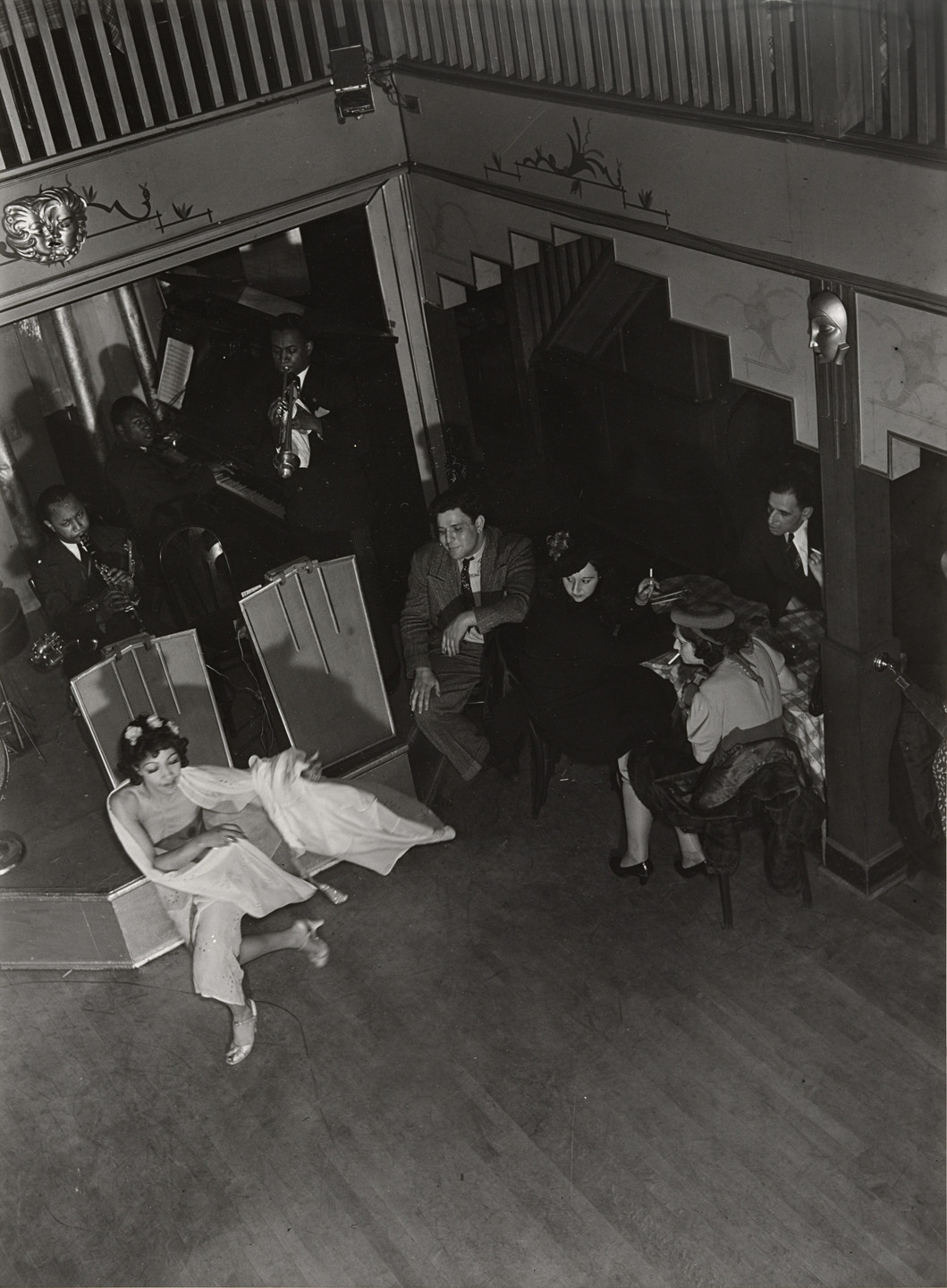 A black-and-white image shows a group of musicians in a corner with audience members seated nearby. In the foreground, someone in a light dress is dancing or performing on the floor. This is an AI generated description. To give feedback, email ai@moma.org.