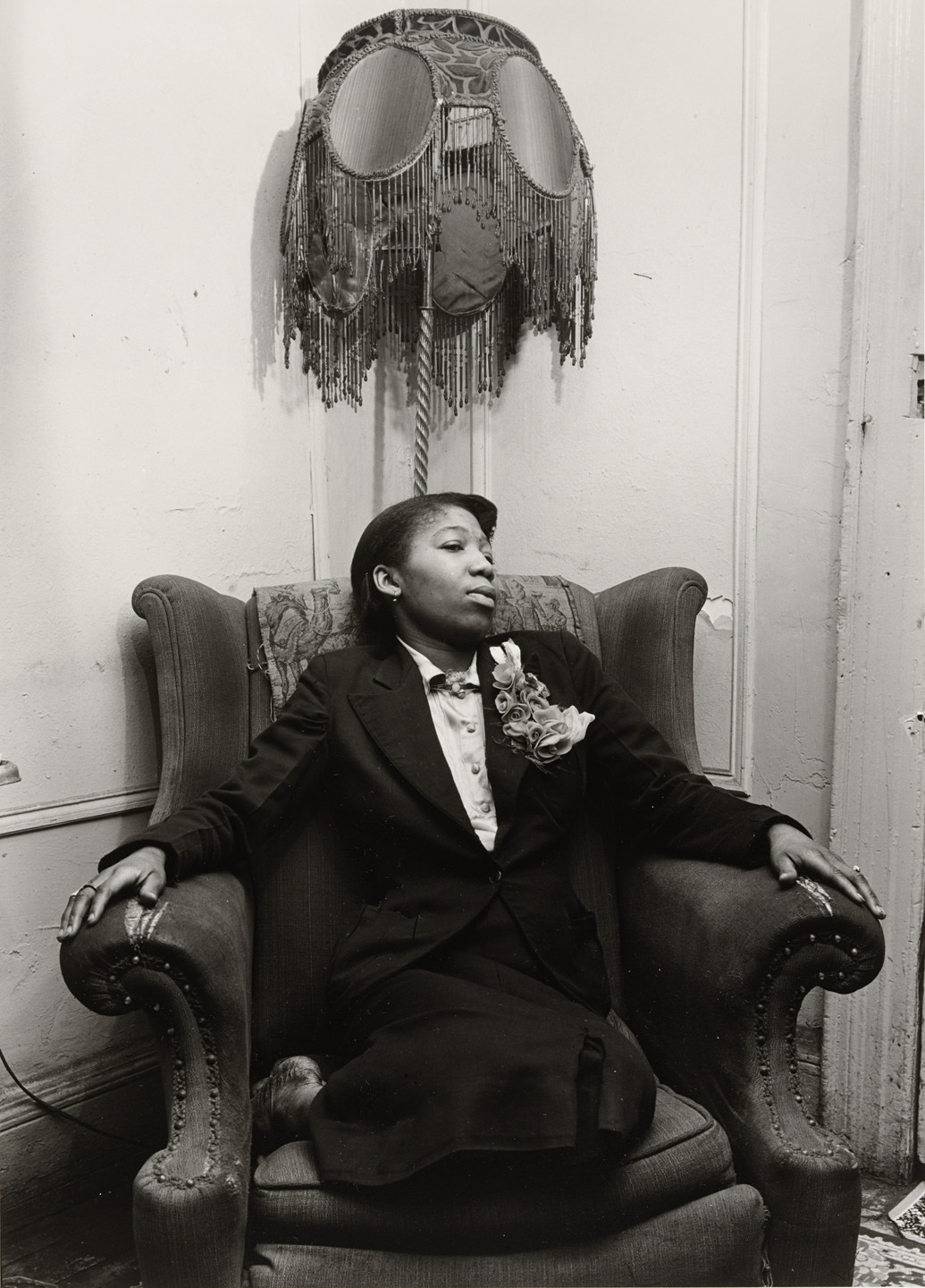 A person in a suit sits in a large armchair with arms resting on the armrests. A fringed lampshade hangs on the wall above the person's head. This is an AI generated description. To give feedback, email ai@moma.org.