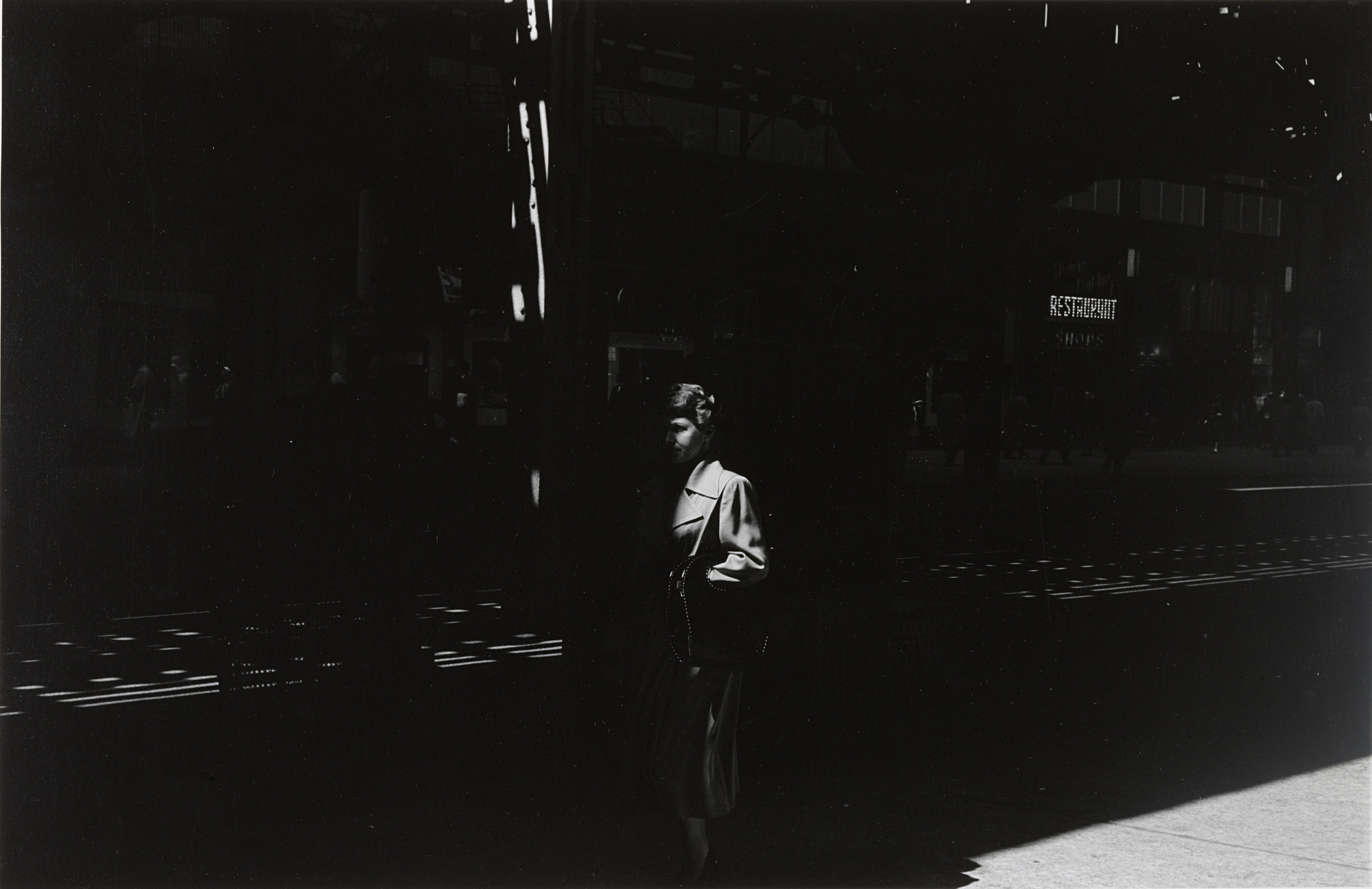 A person wearing a light-colored coat stands under a shadowy structure with beams of light creating streaks across the scene. The surrounding environment is mostly dark with a sign in the distance. This is an AI generated description. To give feedback, email ai@moma.org.