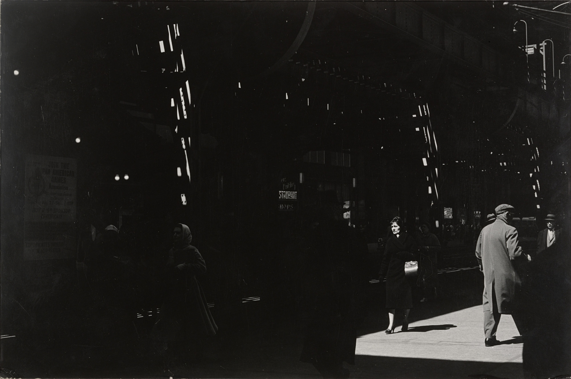 The image shows a dark street scene with a few people walking, primarily illuminated by small beams of light and shadow patterns. Predominant colors are shades of black and grey, with light patches highlighting individuals and parts of the street. This is an AI generated description. To give feedback, email ai@moma.org.