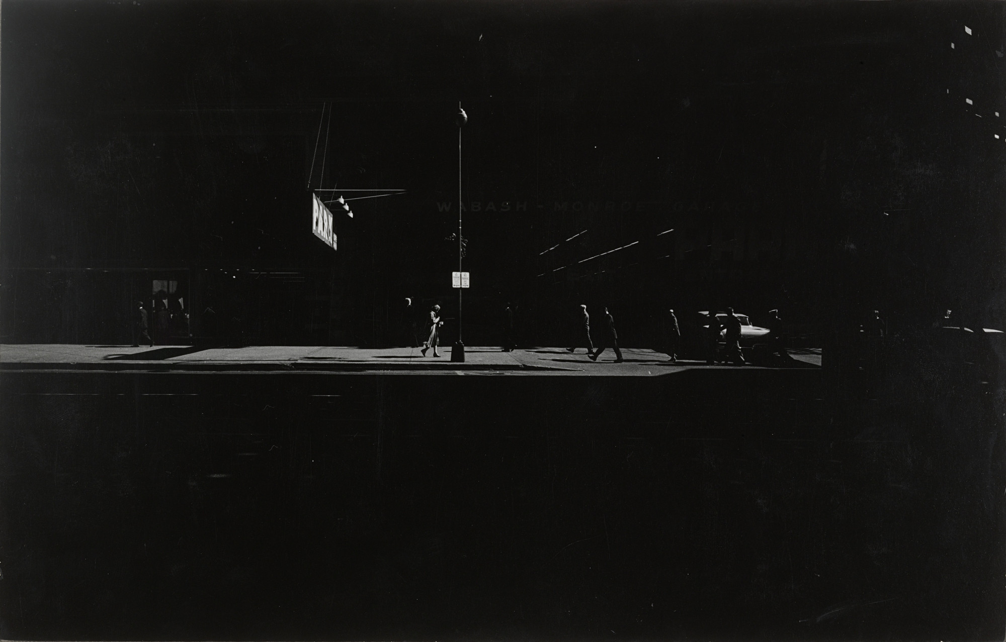 The image shows a dark street scene with a few people walking on a brightly lit sidewalk under a streetlamp. The predominant colors are black and shades of grey with some light areas where the street and figures are illuminated. This is an AI generated description. To give feedback, email ai@moma.org.