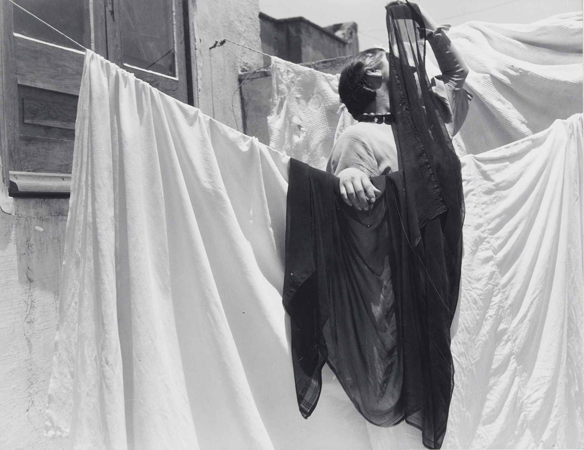 A person stands amongst hanging laundry, holding a semi-transparent dark cloth over their head and body. The predominant colors are white and dark, with visible textures on the fabrics. This is an AI generated description. To give feedback, email ai@moma.org.