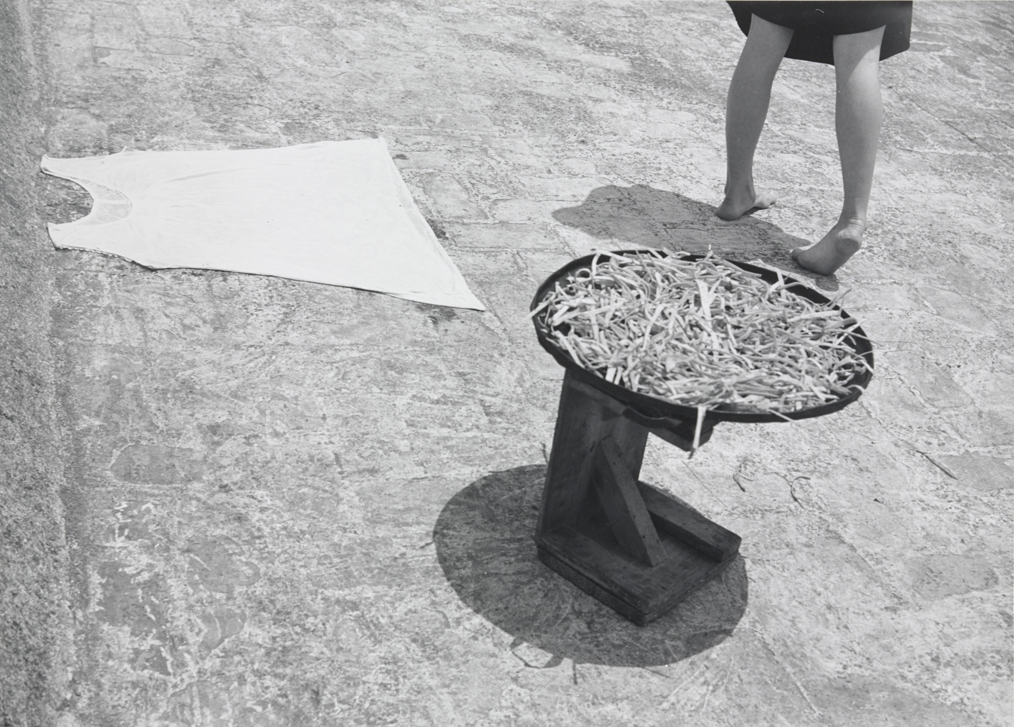 A white sleeveless shirt lies flat on a textured stone floor. Next to it, a person stands barefoot with legs visible, adjacent to a small wooden table holding a bowl of twigs. This is an AI generated description. To give feedback, email ai@moma.org.