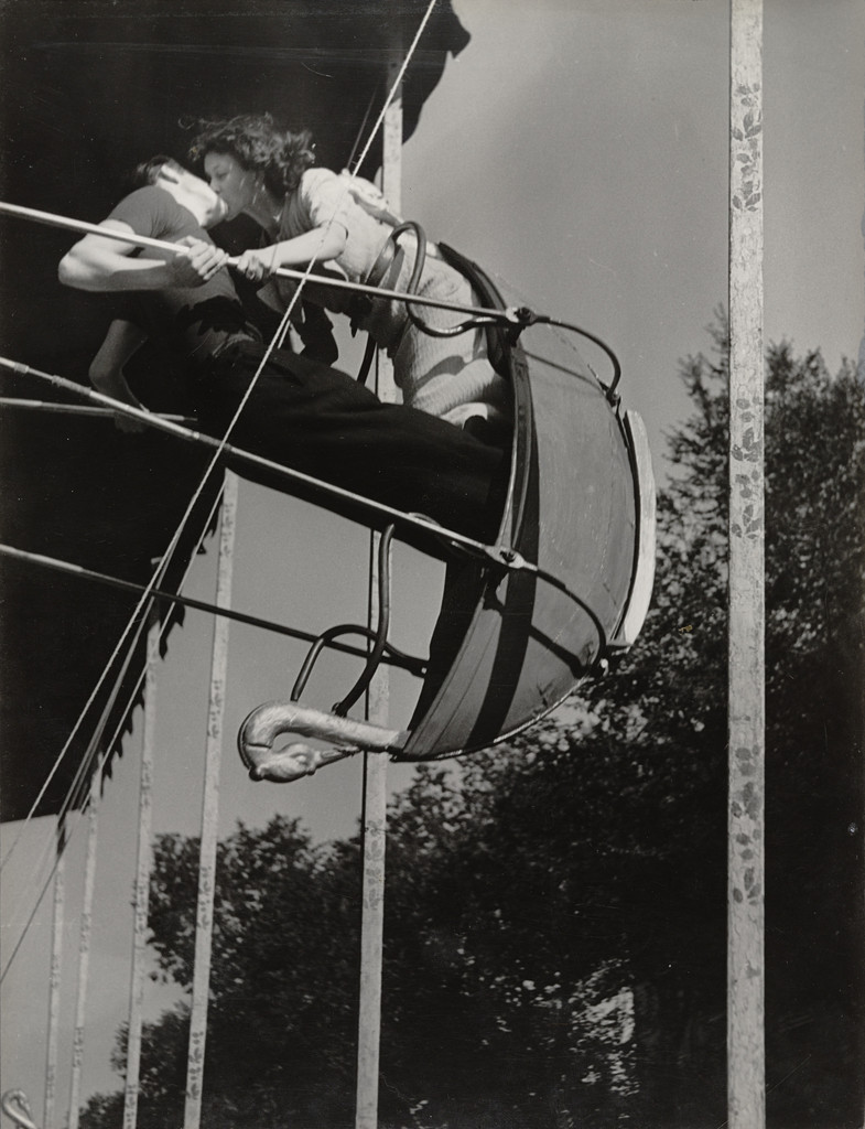A black-and-white photograph by Brassaï showing two people kissing on a fairground swing ride, suspended in mid-air. The swing's metal frame cuts dramatic diagonals across the composition. Their faces are pressed together, oblivious to the camera and the world below.