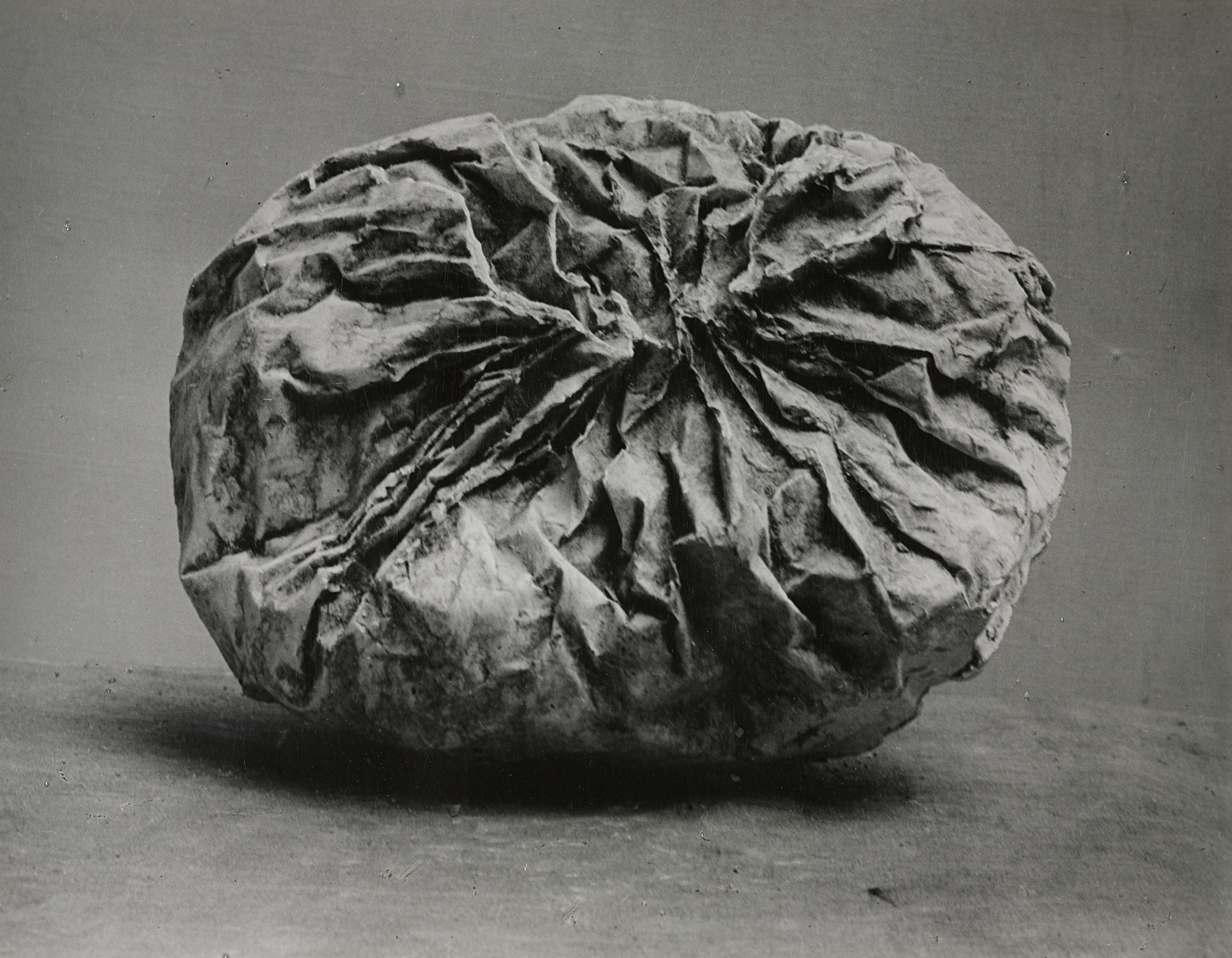 A photograph shows a crumpled, spherical object with deep creases and folds on its surface. The image is predominantly gray, highlighting the texture and details of the object&#39;s rugged surface. This is an AI generated description. To give feedback, email ai@moma.org.