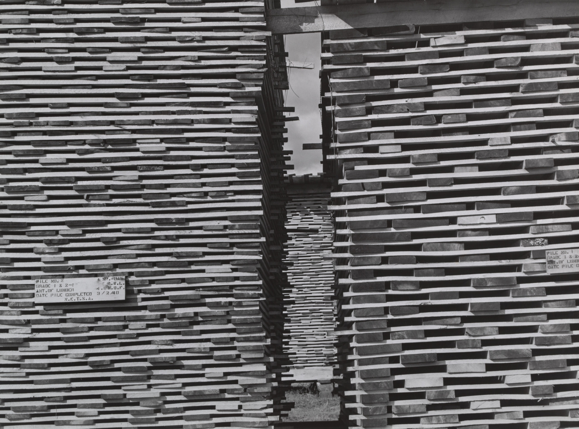 The image shows stacks of cut wooden planks piled horizontally with a narrow gap between two large stacks. The predominant colors are shades of gray and brown, creating a layered pattern effect. This is an AI generated description. To give feedback, email ai@moma.org.