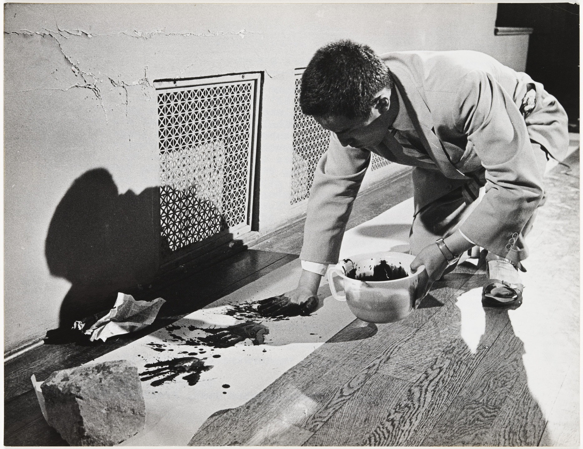 A person in a suit is kneeling on the floor, holding a bowl of dark liquid, and painting on a large white sheet of paper. The scene is in black and white, with a textured wall and vent in the background. This is an AI generated description. To give feedback, email ai@moma.org.