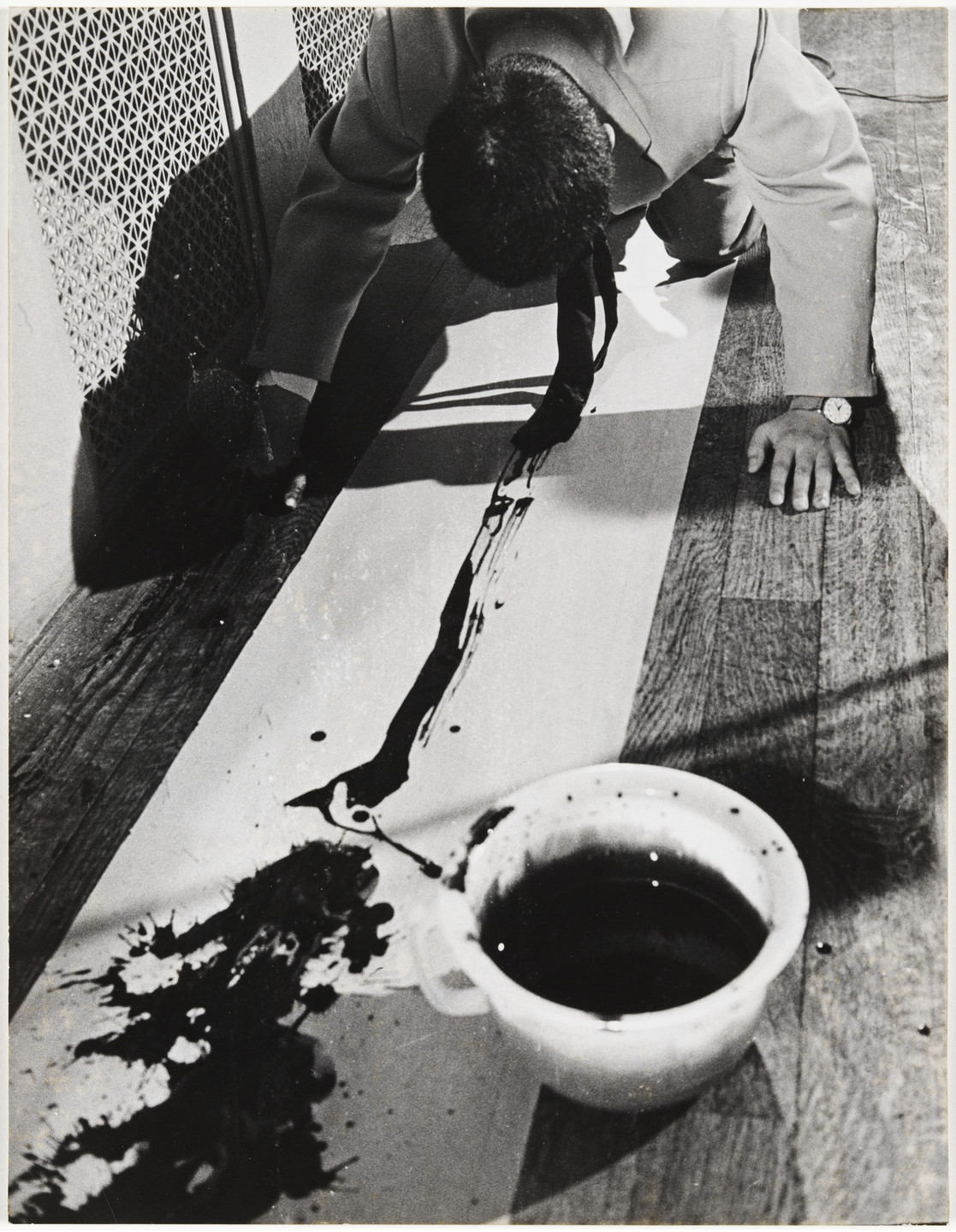 A person in a light-colored suit kneels on a wooden floor, drawing a black line across a large white surface with a paintbrush. A white cup filled with dark liquid sits nearby, with splatters surrounding it. This is an AI generated description. To give feedback, email ai@moma.org.