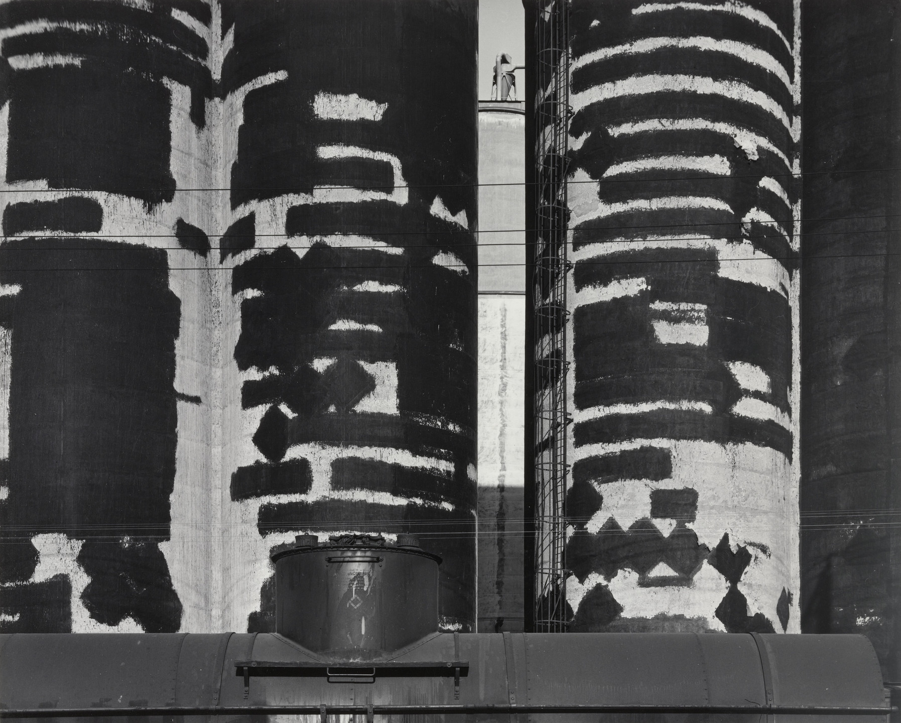 The image shows very tall, cylindrical structures with black and white, irregular patterns. A small figure stands at the top of a ladder between the structures. This is an AI generated description. To give feedback, email ai@moma.org.