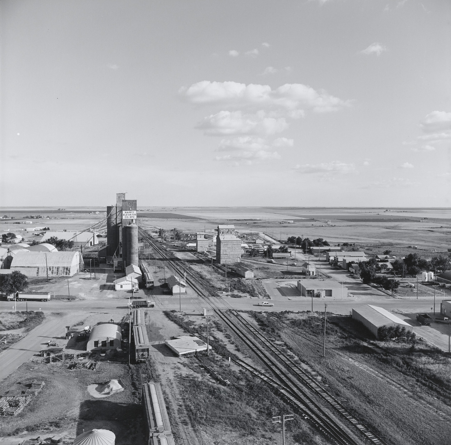 A black-and-white aerial view of a rural town shows railroad tracks running from the foreground to the horizon, flanked by grain elevators and small buildings. The sky is partly cloudy, and expansive fields stretch to the horizon. This is an AI generated description. To give feedback, email ai@moma.org.