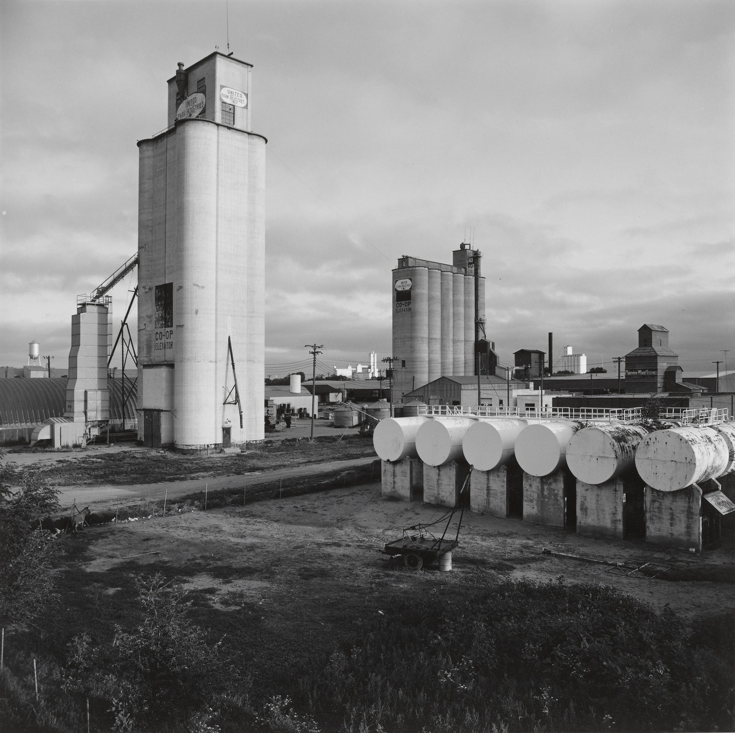 The image features an industrial landscape with several tall, cylindrical storage silos and horizontal tanks in the foreground, all in grayscale tones. There are buildings and equipment spread throughout the scene, with a cloudy sky overhead. This is an AI generated description. To give feedback, email ai@moma.org.
