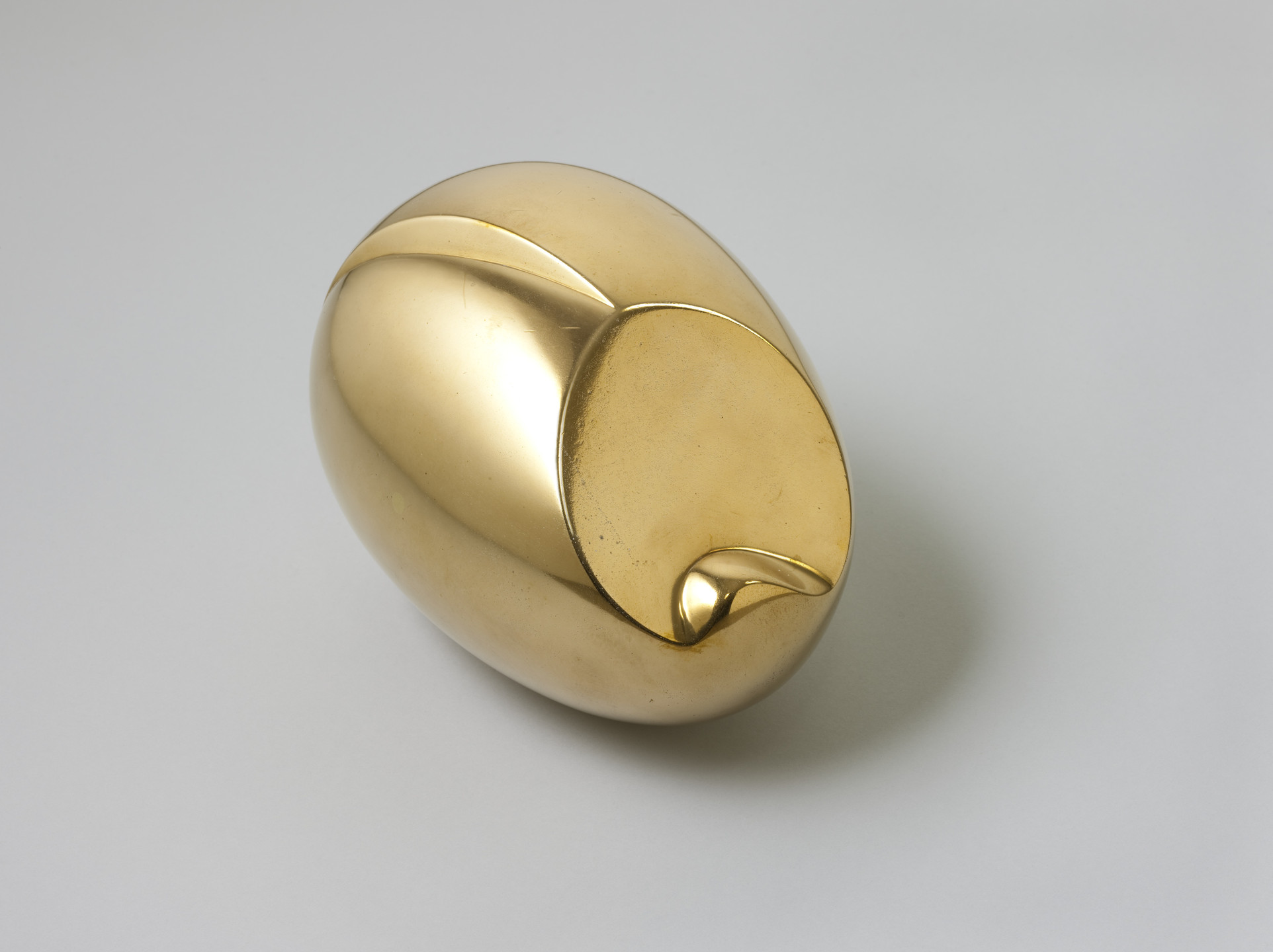 A smooth, golden, egg-shaped sculpture with a shiny surface is lying on its side against a light gray background. It features a small, pointed protrusion and curved indentations. This is an AI generated description. To give feedback, email ai@moma.org.