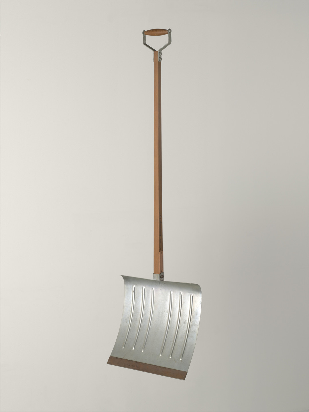 A snow shovel with a long wooden handle and a metallic blade is centered against a plain, light gray background. The shovel blade is silver with vertical grooves and a slightly curved shape. This is an AI generated description. To give feedback, email ai@moma.org.