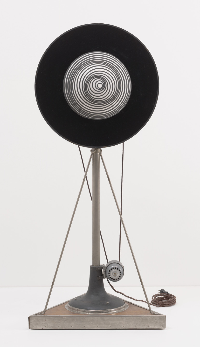A metallic structure stands on a triangular base with a central circular black disk featuring concentric silver rings. A motor-like device and cords are attached to the structure. This is an AI generated description. To give feedback, email ai@moma.org.