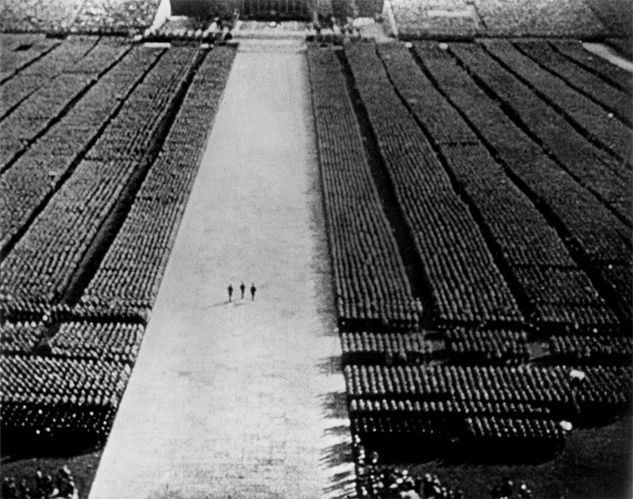 A large gathering of people is seen from above, standing in structured rows on both sides of a wide central pathway. Three individuals walk in the center of this pathway, moving towards the top of the image. This is an AI generated description. To give feedback, email ai@moma.org.