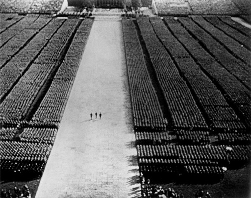 A large gathering of people is seen from above, standing in structured rows on both sides of a wide central pathway. Three individuals walk in the center of this pathway, moving towards the top of the image. This is an AI generated description. To give feedback, email ai@moma.org.