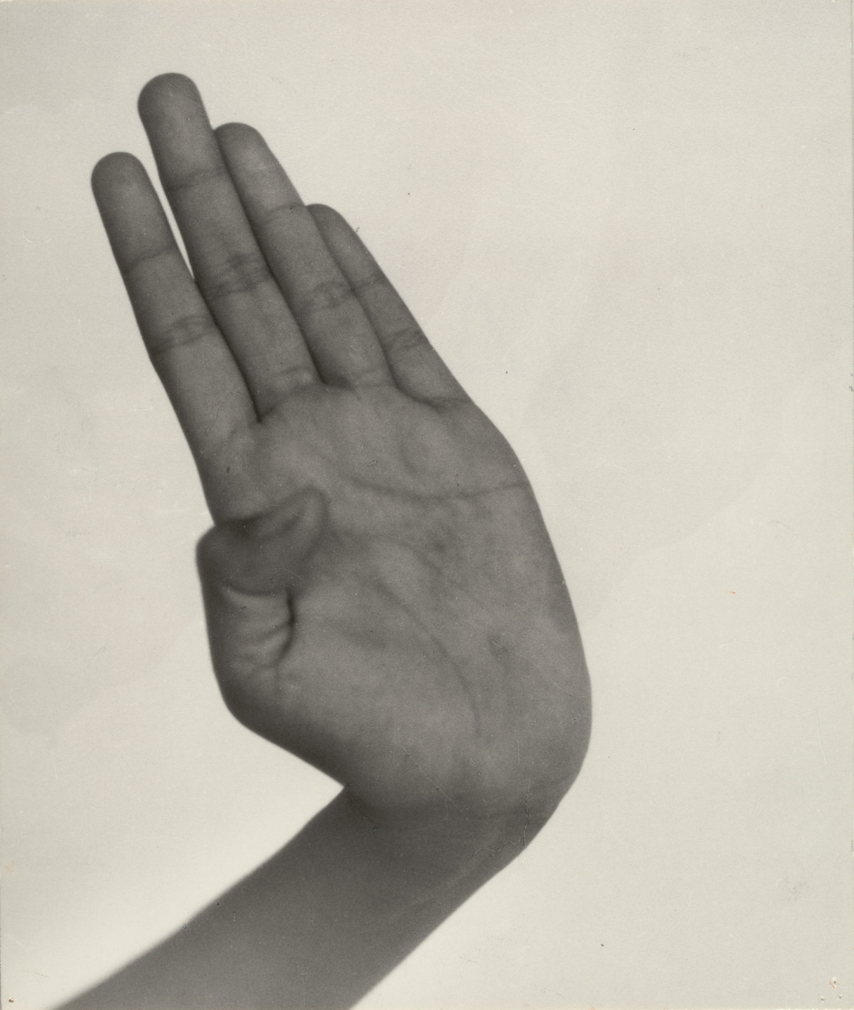 A grayscale image of an open hand stretching upward with fingers together and thumb bent inward. The background is light gray, and the hand is centrally positioned, occupying most of the frame. This is an AI generated description. To give feedback, email ai@moma.org.