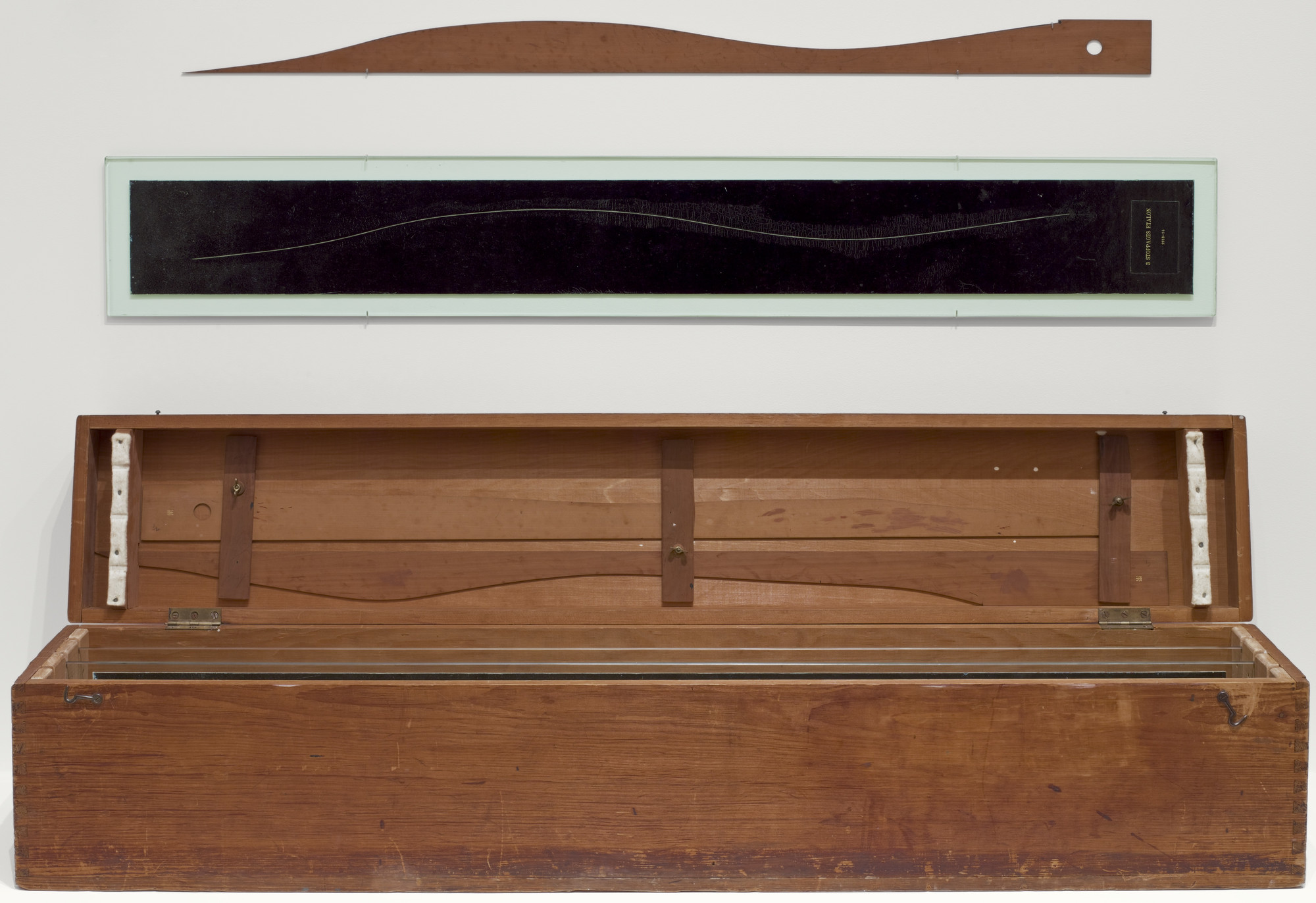 A long, open wooden box with a hinged lid, placed below a horizontal black panel with a wavy line and a curved, wooden tool above it. The predominant colors are brown and black. This is an AI generated description. To give feedback, email ai@moma.org.