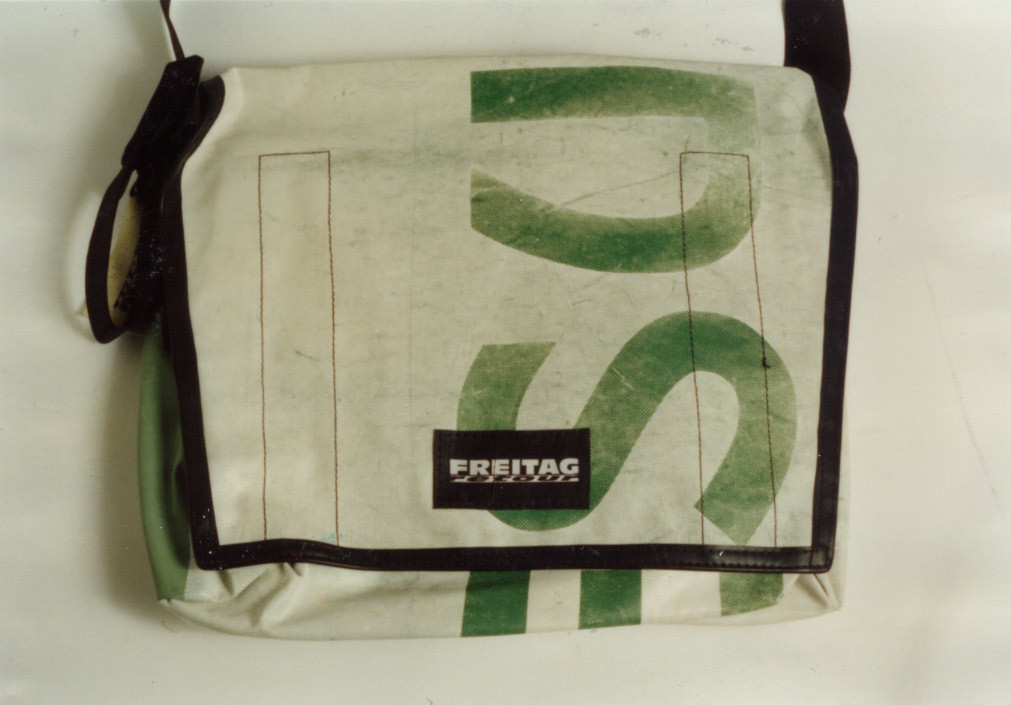 A white messenger bag with black edges and a black strap. The bag has large green letters on its front and a black patch with the word &#34;FREITAG&#34; on a white background. This is an AI generated description. To give feedback, email ai@moma.org.