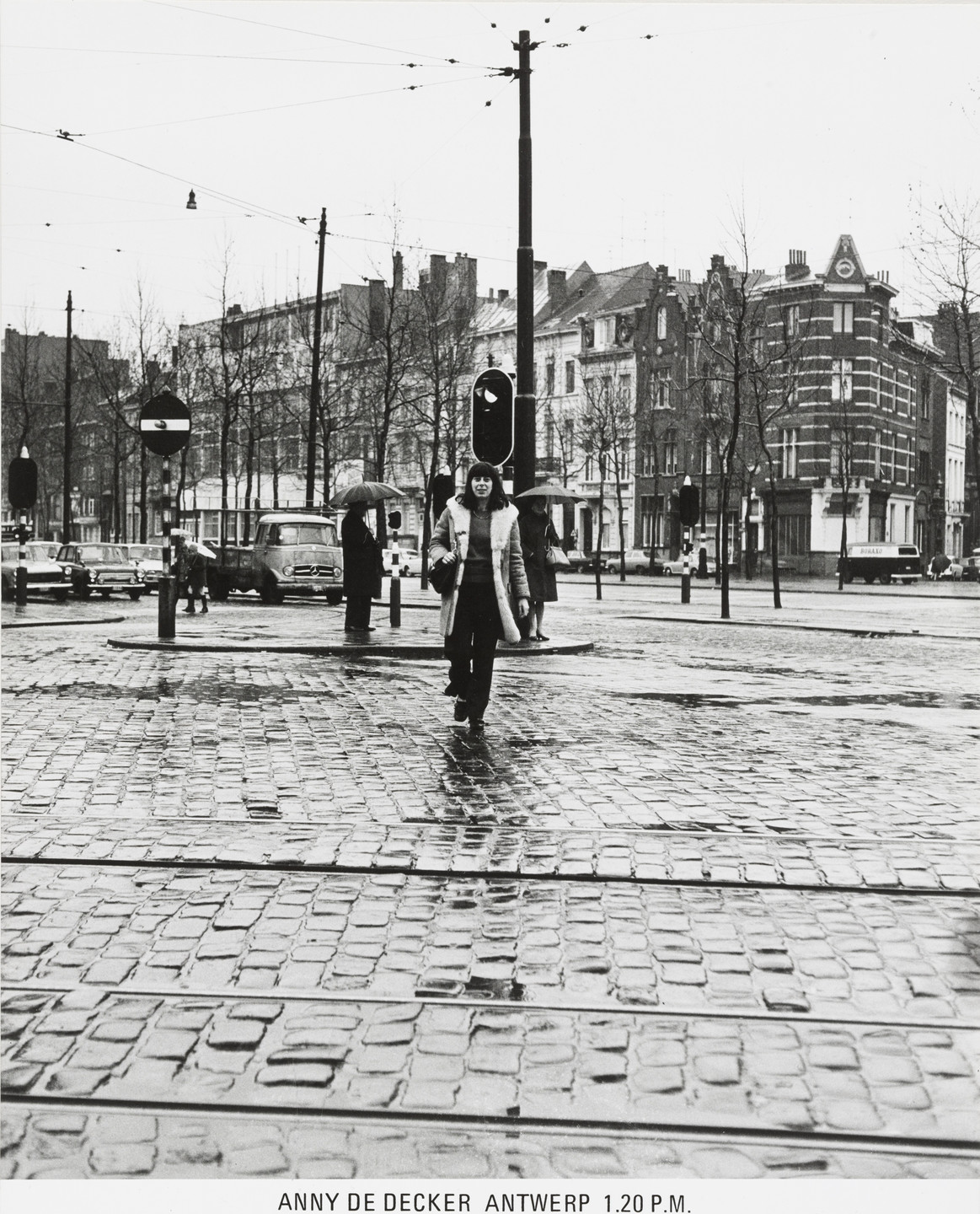 A person walks across a wet cobblestone street in a city with brick buildings and leafless trees; others hold umbrellas. Predominant colors are shades of grey and black. This is an AI generated description. To give feedback, email ai@moma.org.