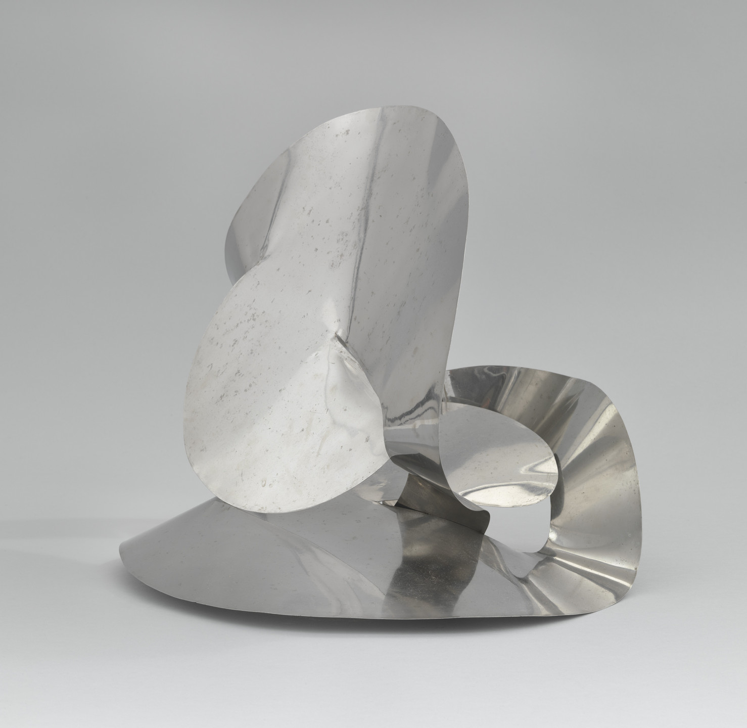 A polished silver metal sculpture with smooth, flowing, and curved surfaces is displayed. The piece reflects light and has organic, asymmetrical shapes that overlap each other. This is an AI generated description. To give feedback, email ai@moma.org.