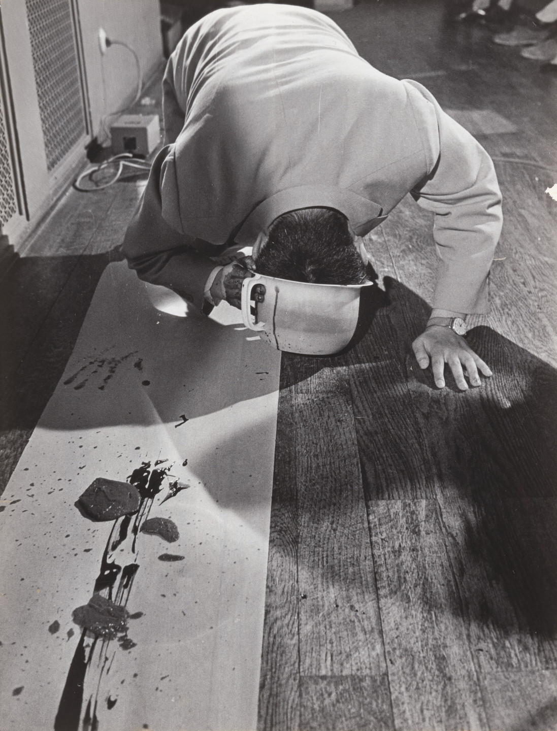 A person in a light-colored suit is kneeling on a wooden floor, their head close to a white canvas with black smudges and objects on it. There is a cup near their face and some cables on the floor. This is an AI generated description. To give feedback, email ai@moma.org.