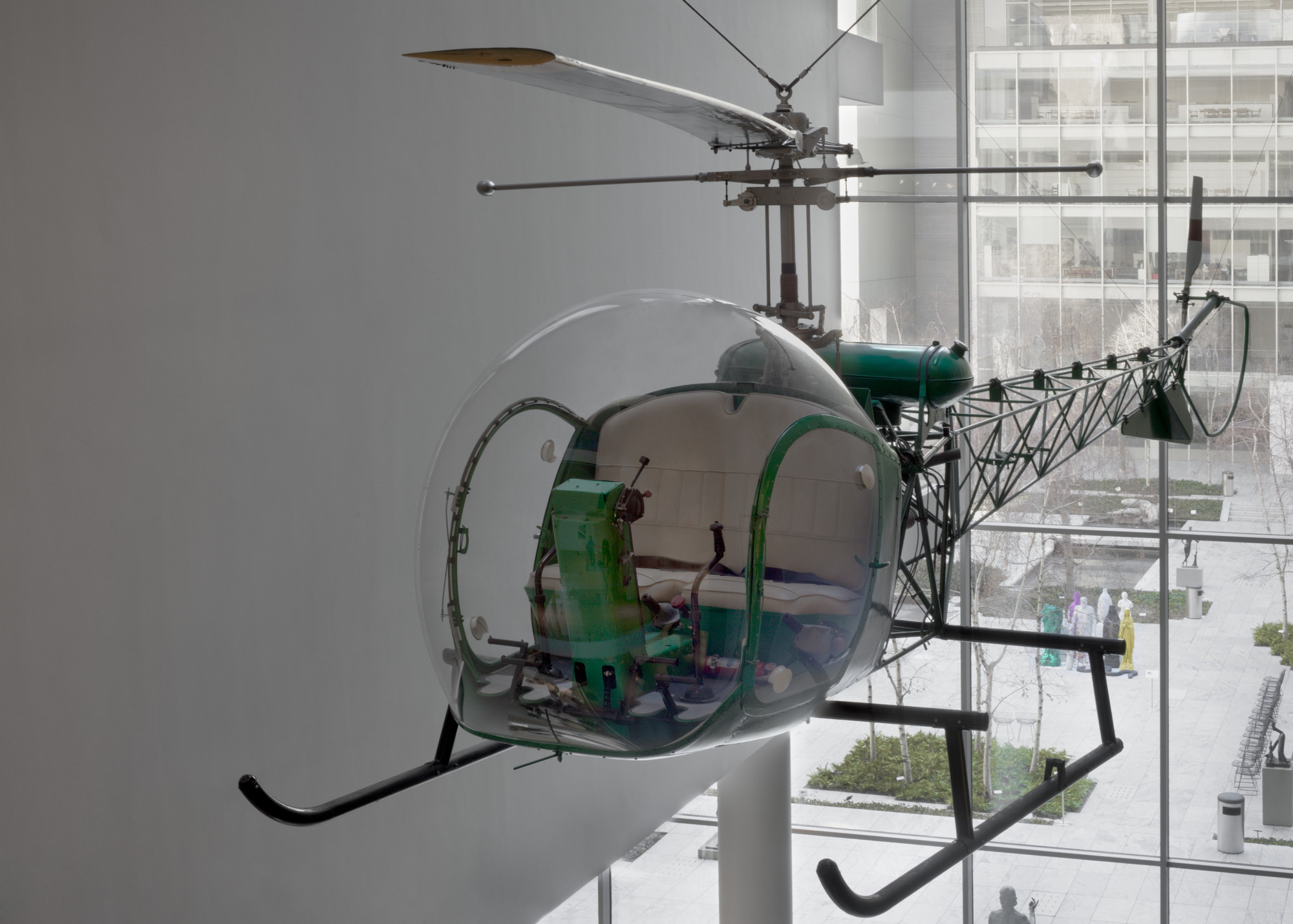 A green helicopter with a clear bubble cockpit, white seat, and black propellor and skids, is suspended indoors. Behind it is a window revealing an outdoor courtyard with trees and modern buildings. This is an AI generated description. To give feedback, email ai@moma.org.