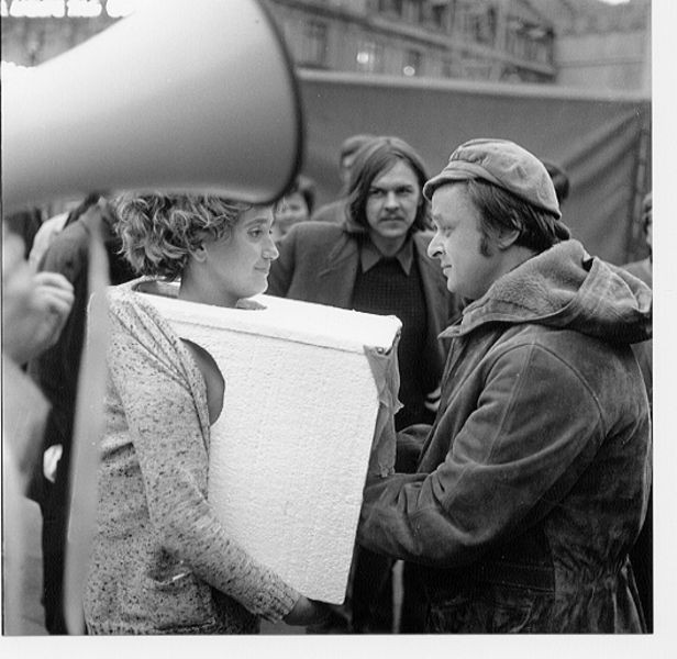 A black-and-white image shows two people facing each other; one holds a large rectangular object. An out-of-focus figure is in the background, and a large horn is visible in the foreground. This is an AI generated description. To give feedback, email ai@moma.org.