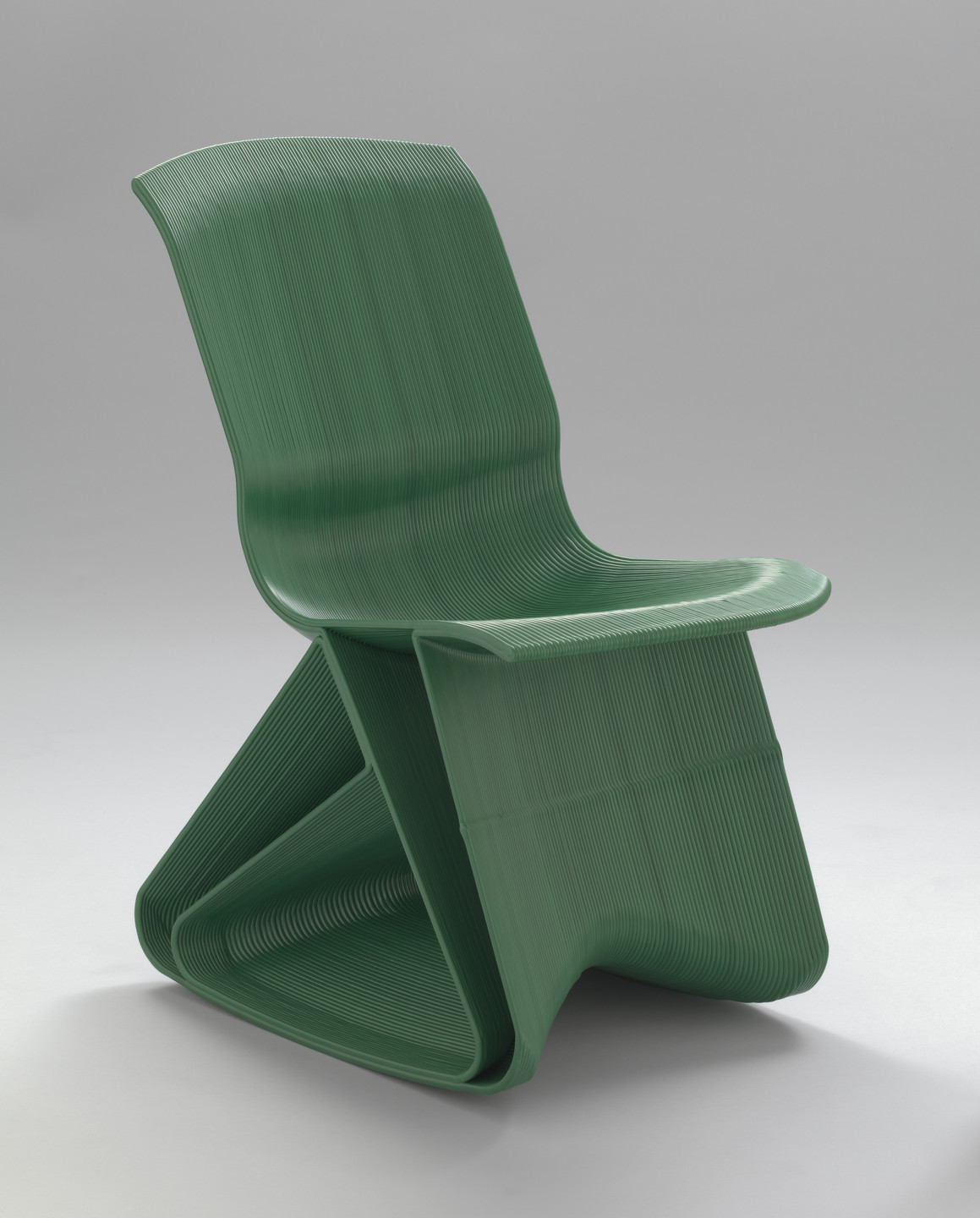 A green chair with a unique, continuous loop design sits on a plain, light gray background. The chair has a ribbed texture and a geometric, folded shape. This is an AI generated description. To give feedback, email ai@moma.org.