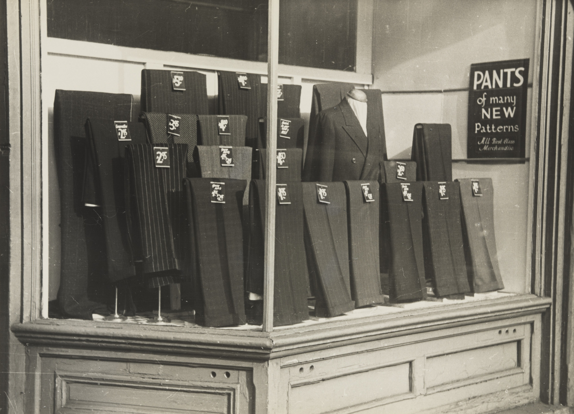 A store window display showcases folded pants in various dark shades like black and gray with price tags, alongside a suit jacket on a mannequin. A sign reads &#34;PANTS of many NEW Patterns, All First Class Merchandise.&#34; This is an AI generated description. To give feedback, email ai@moma.org.