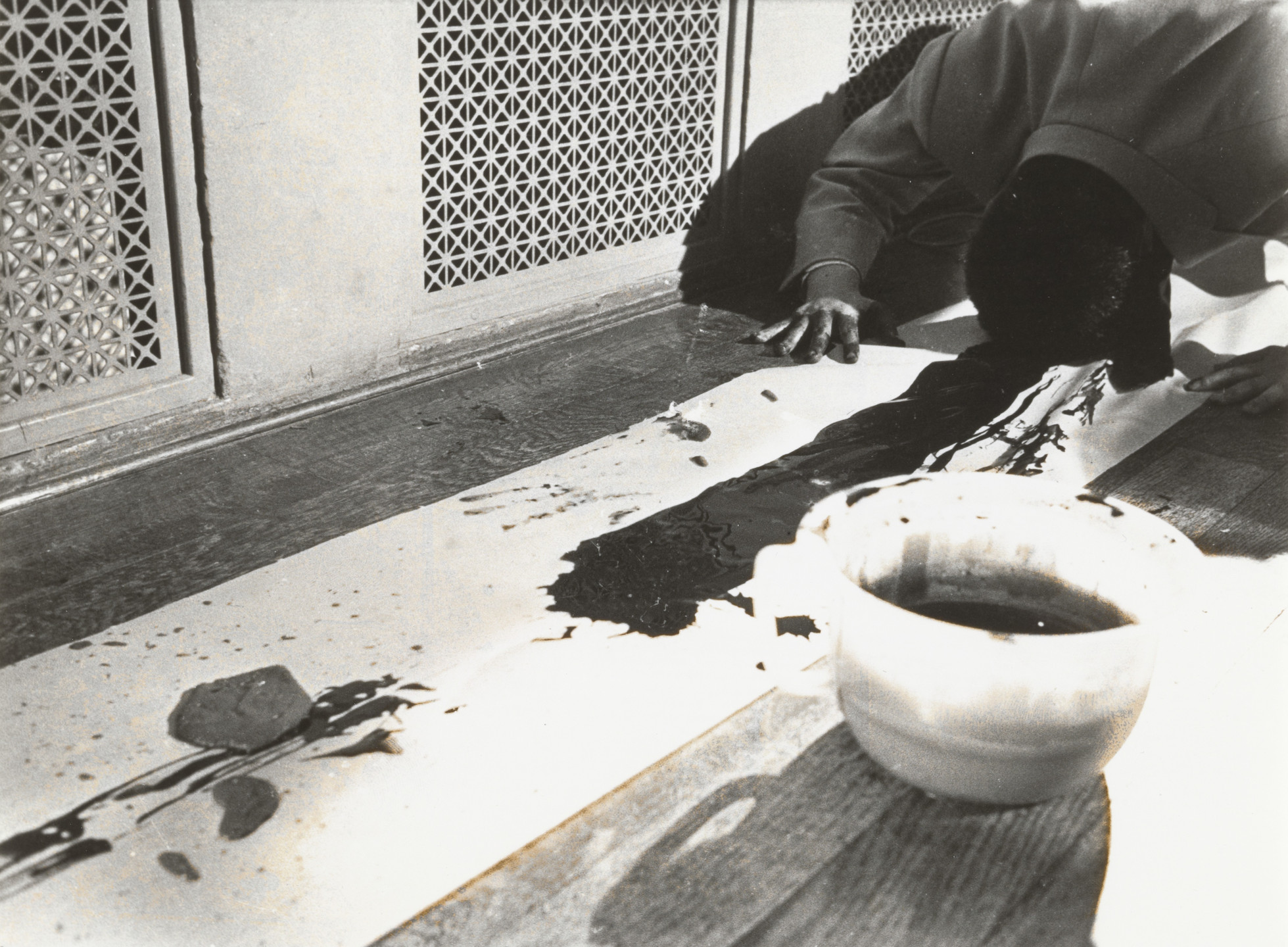 A person is on the floor, leaning over a long white sheet with black paint on it, next to a white bowl containing dark liquid. The background features a patterned wall with a lattice design. This is an AI generated description. To give feedback, email ai@moma.org.