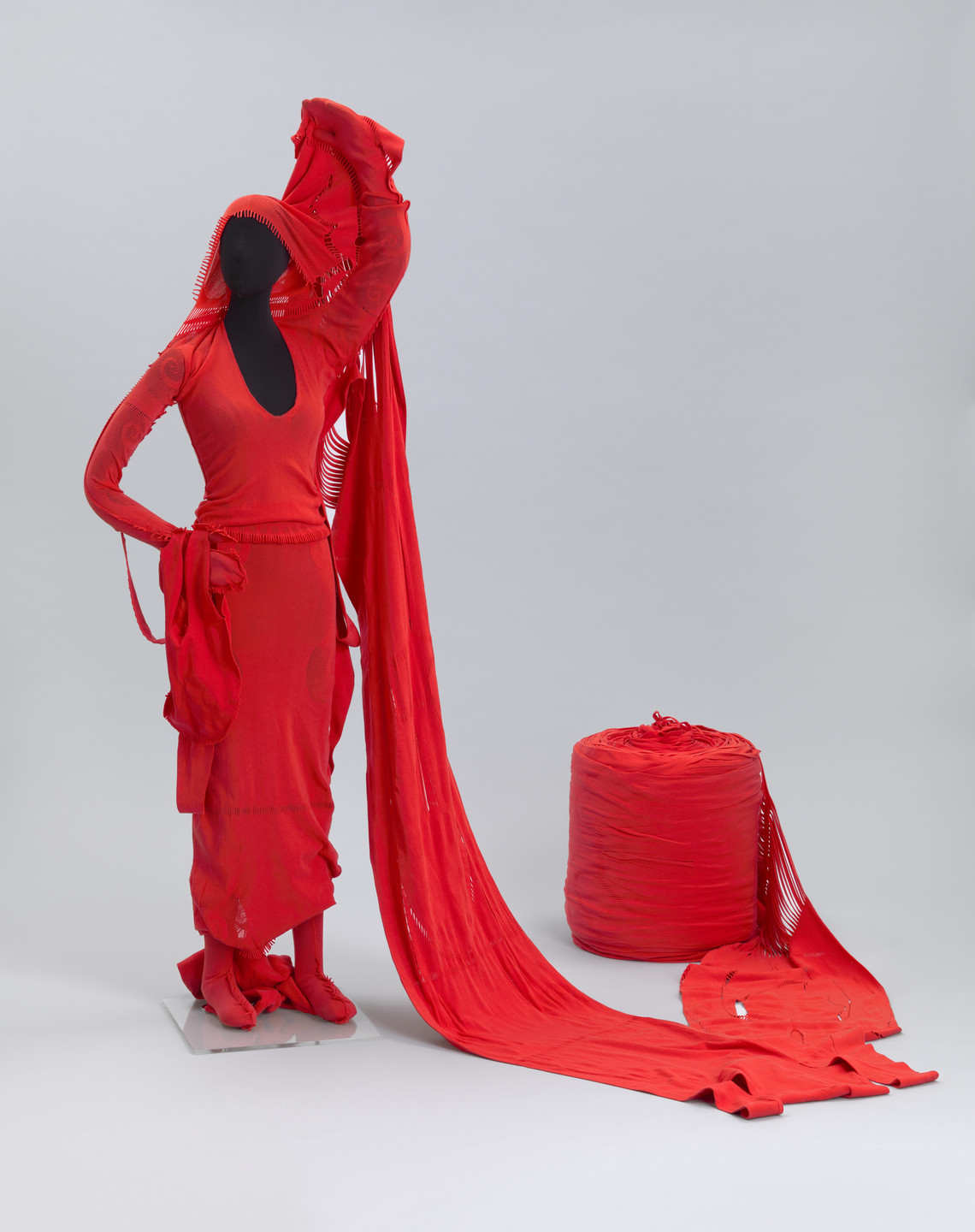 A black mannequin dressed in a red outfit stands with one arm raised, holding a long red fabric that extends to the floor. A cylindrical bundle of red fabric is placed on the ground beside the mannequin. This is an AI generated description. To give feedback, email ai@moma.org.