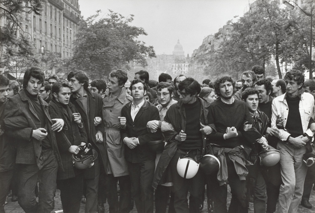 A horizontal line of young demonstrators linking arms in the middle of a Paris street. Men and women, leather jackets and trench coats, serious faces. Behind them, the avenue stretches back toward trees and classical buildings. In the far distance, barely visible through the haze, the dome of the Panthéon. Henri Cartier-Bresson's decisive moment.