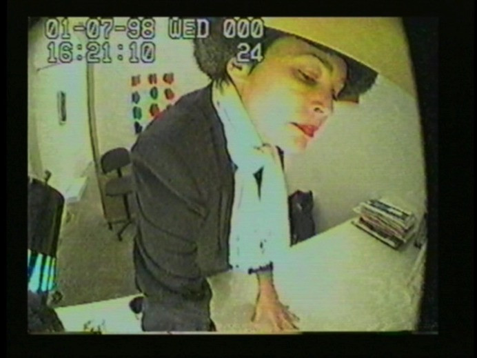 A person in a black outfit and white scarf is leaning over a counter, looking down, with their hand resting on the surface. The background includes stacked papers and a wall decorated with colorful shapes. This is an AI generated description. To give feedback, email ai@moma.org.