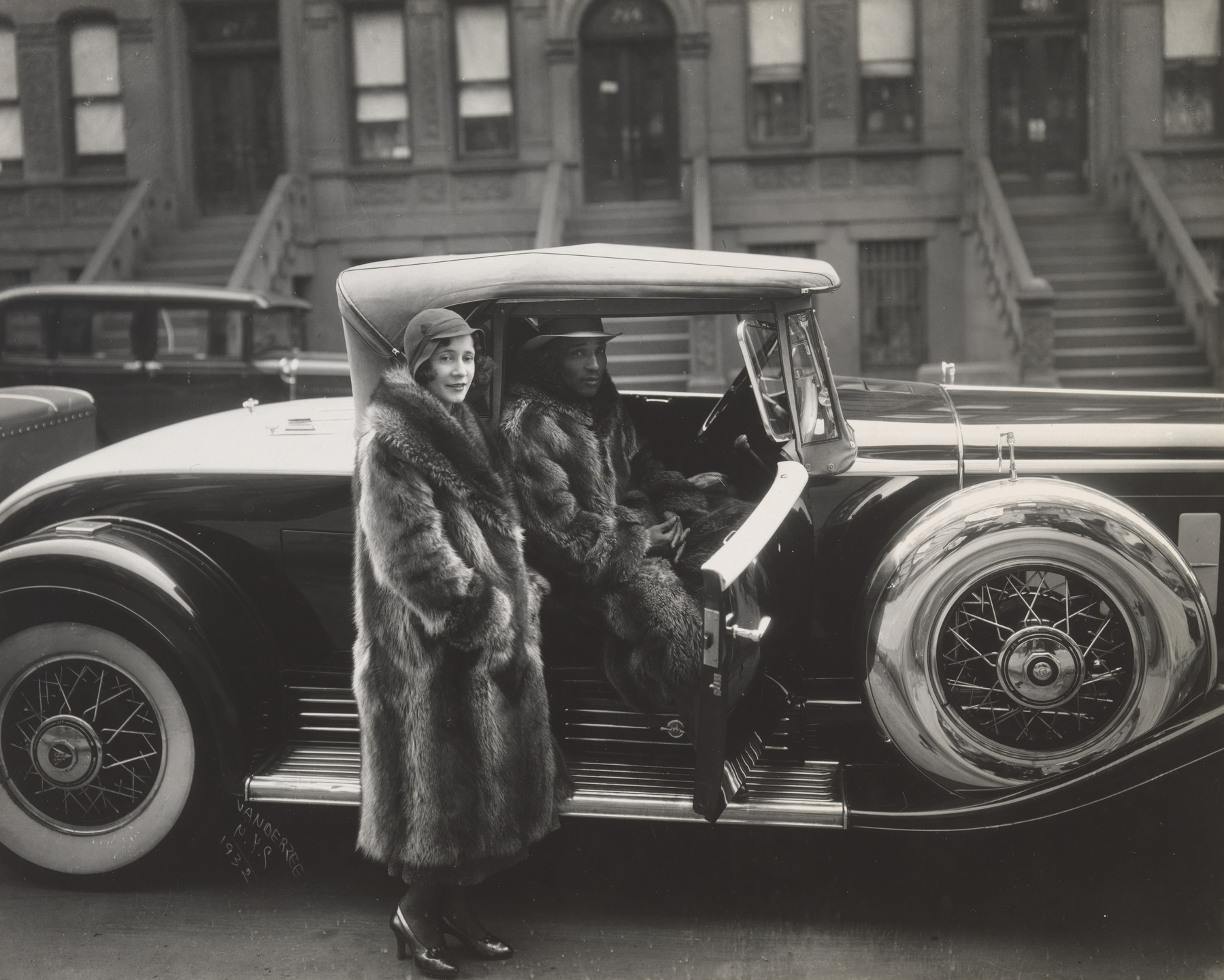Two people in fur coats stand beside a vintage car, with one person in the driver's seat and the other standing outside. The background shows a row of brownstone buildings with stoops. This is an AI generated description. To give feedback, email ai@moma.org.