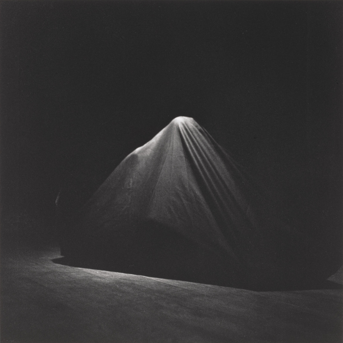 The image shows a dark scene with a draped cloth covering a mound-like shape, primarily in shades of black and gray. The cloth is illuminated from above, creating soft highlights on its folds. This is an AI generated description. To give feedback, email ai@moma.org.