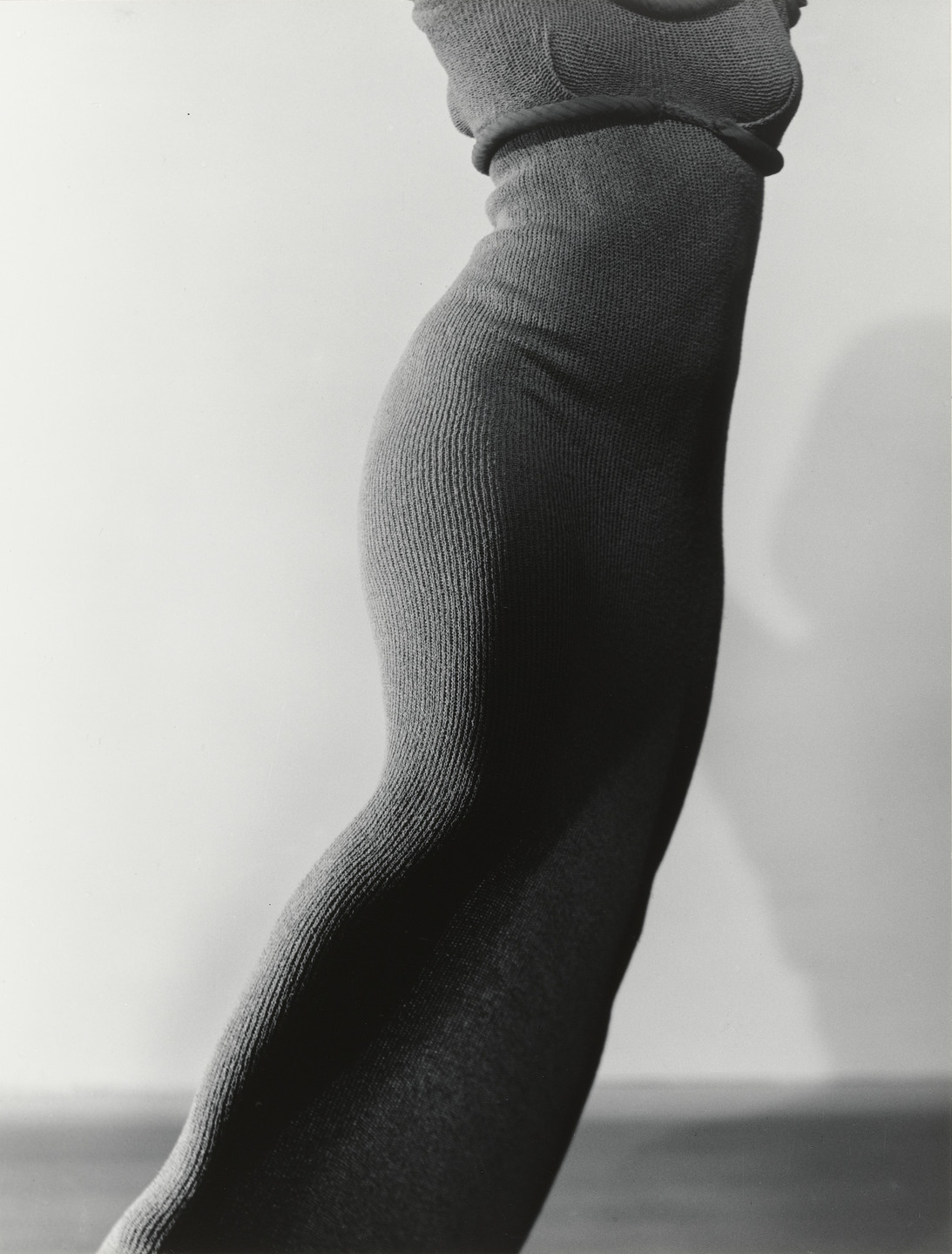 The image shows a close-up of a figure, covered in a form-fitting, textured, dark grey garment, against a light grey background. The figure is depicted in a mid-movement pose emphasizing its curves. This is an AI generated description. To give feedback, email ai@moma.org.