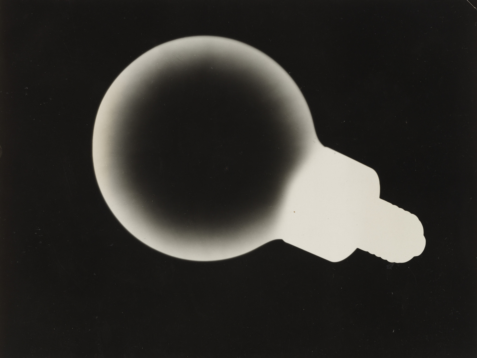 The image shows a black background with the silhouette of a round object, possibly a lightbulb, transitioning from dark to light shades of gray and white. The object&#39;s base appears textured and tapering. This is an AI generated description. To give feedback, email ai@moma.org.